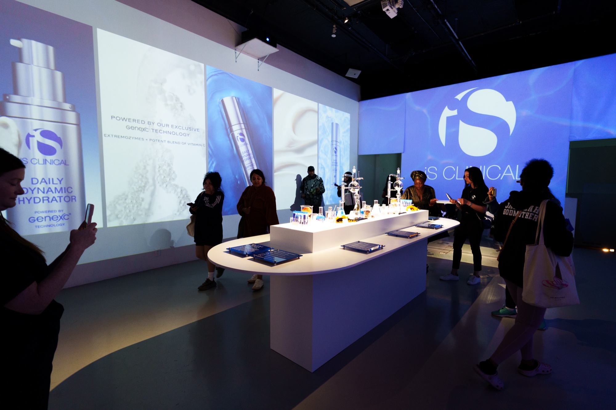Guests photograph a white product table under blue iS Clinical projections and a giant silver bottle graphic