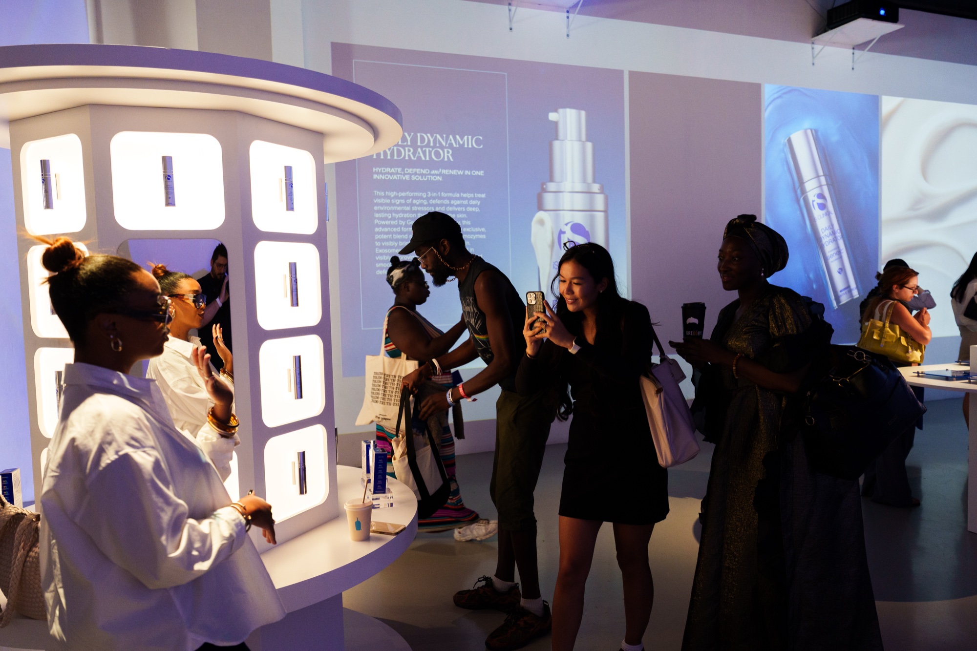 Guests photograph and browse a backlit column of serum bottles under a projected Daily Dynamic Hydrator ad