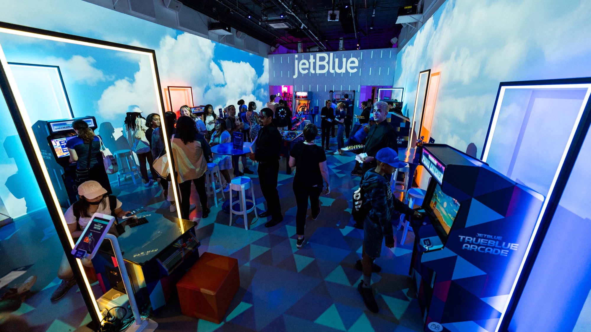 Guests play arcade cabinets under a JetBlue sign in a room projected with blue sky and clouds