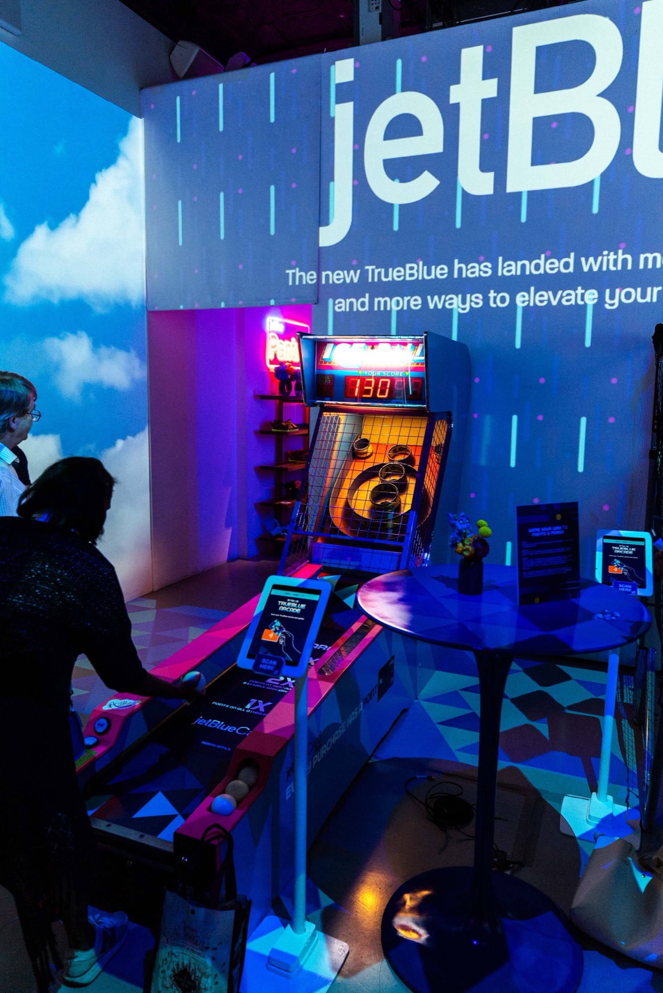 Guest playing a skee ball lane beneath a giant jetBlue projection and a cloud wall in blue light