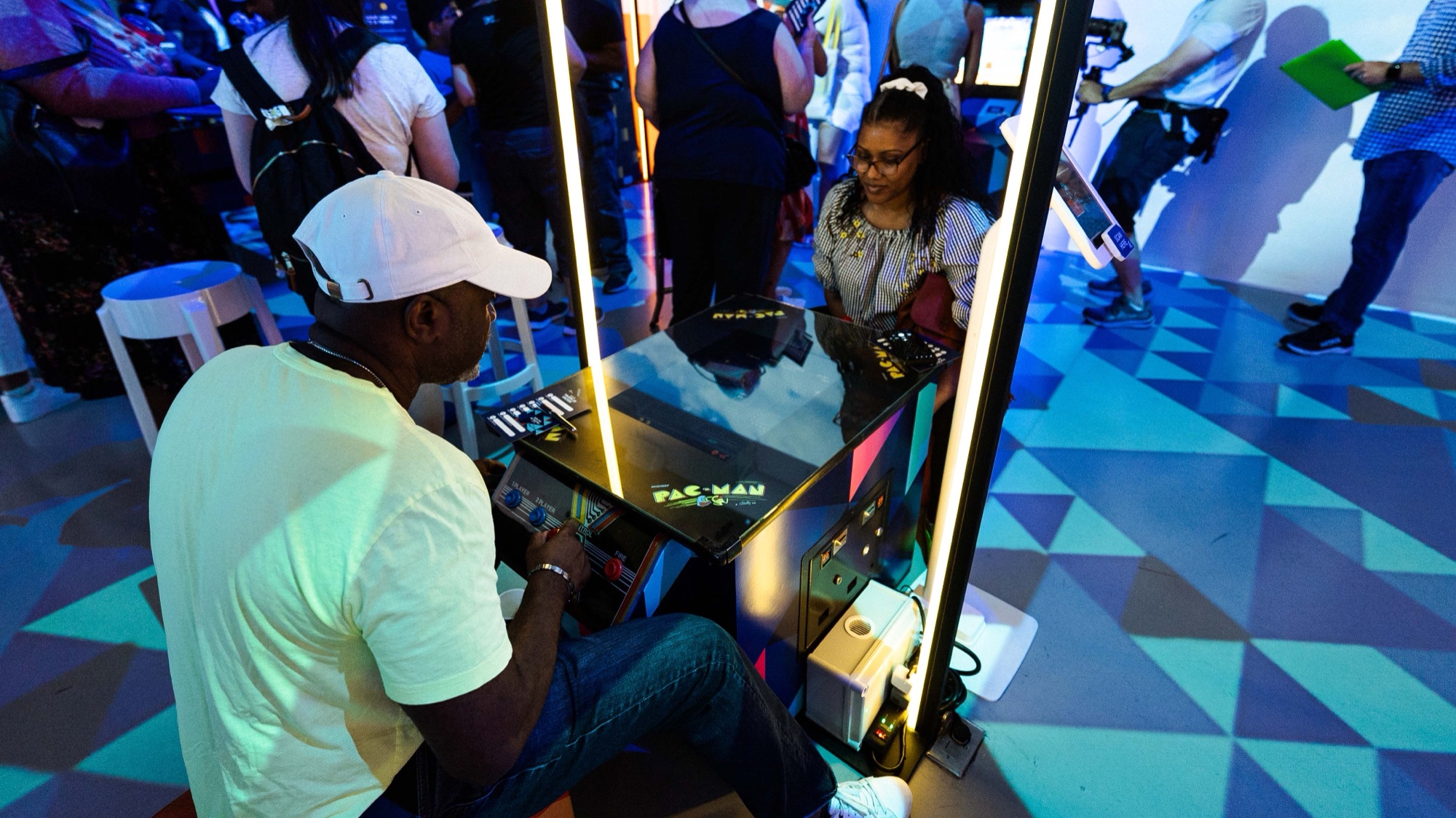 Man in a white cap playing a Pac Man arcade table opposite a woman on a blue geometric floor