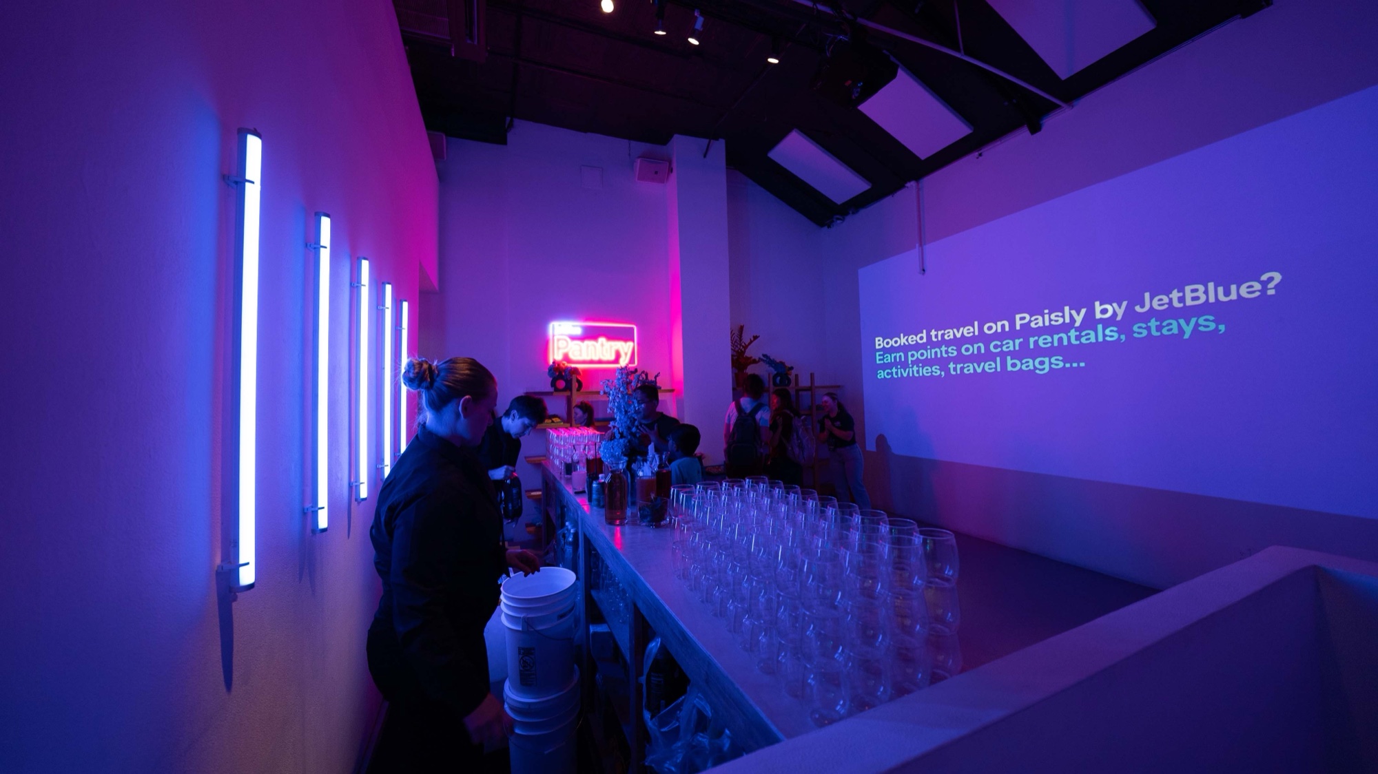Rows of empty glasses line a white bar in a blue and pink room with JetBlue text on the wall