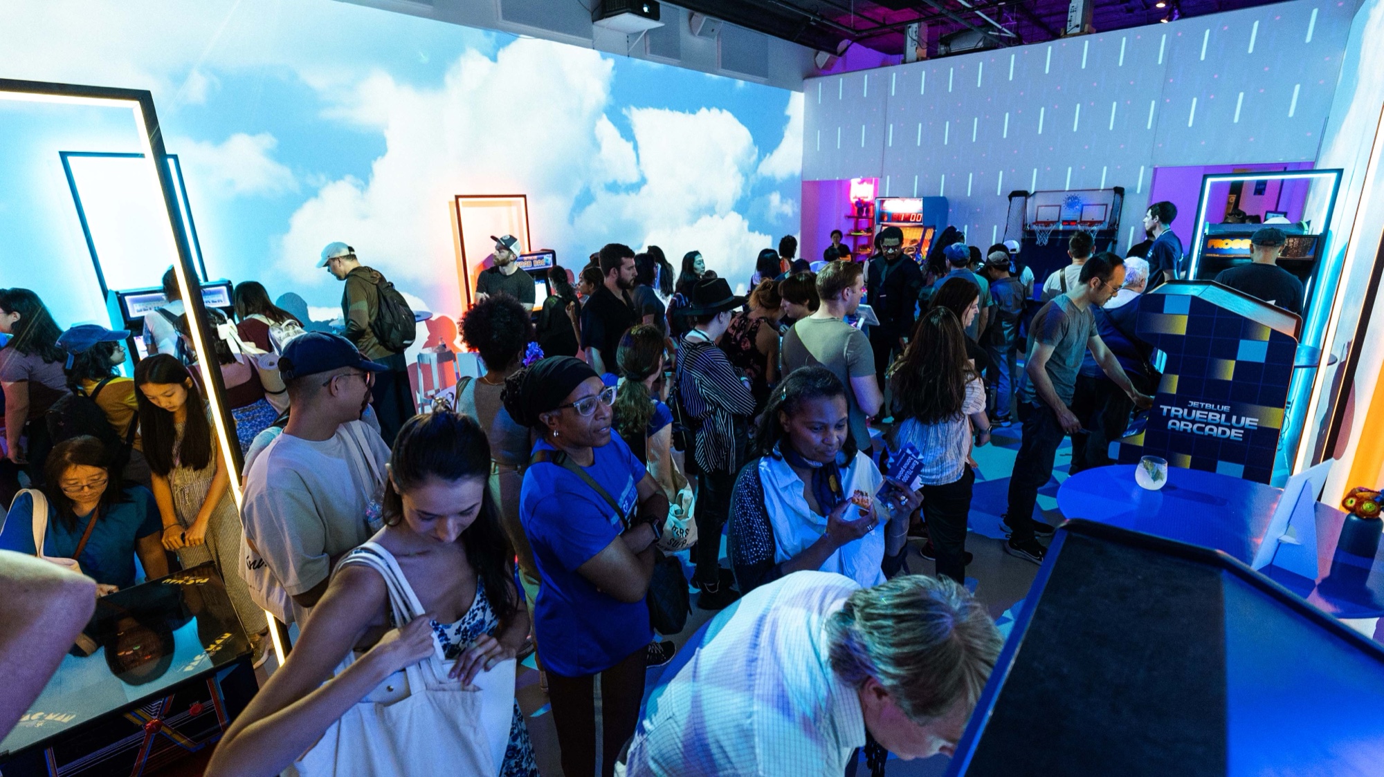 Guests fill a blue lit room with a projected cloud sky, glowing mirror frames and a pixel style display