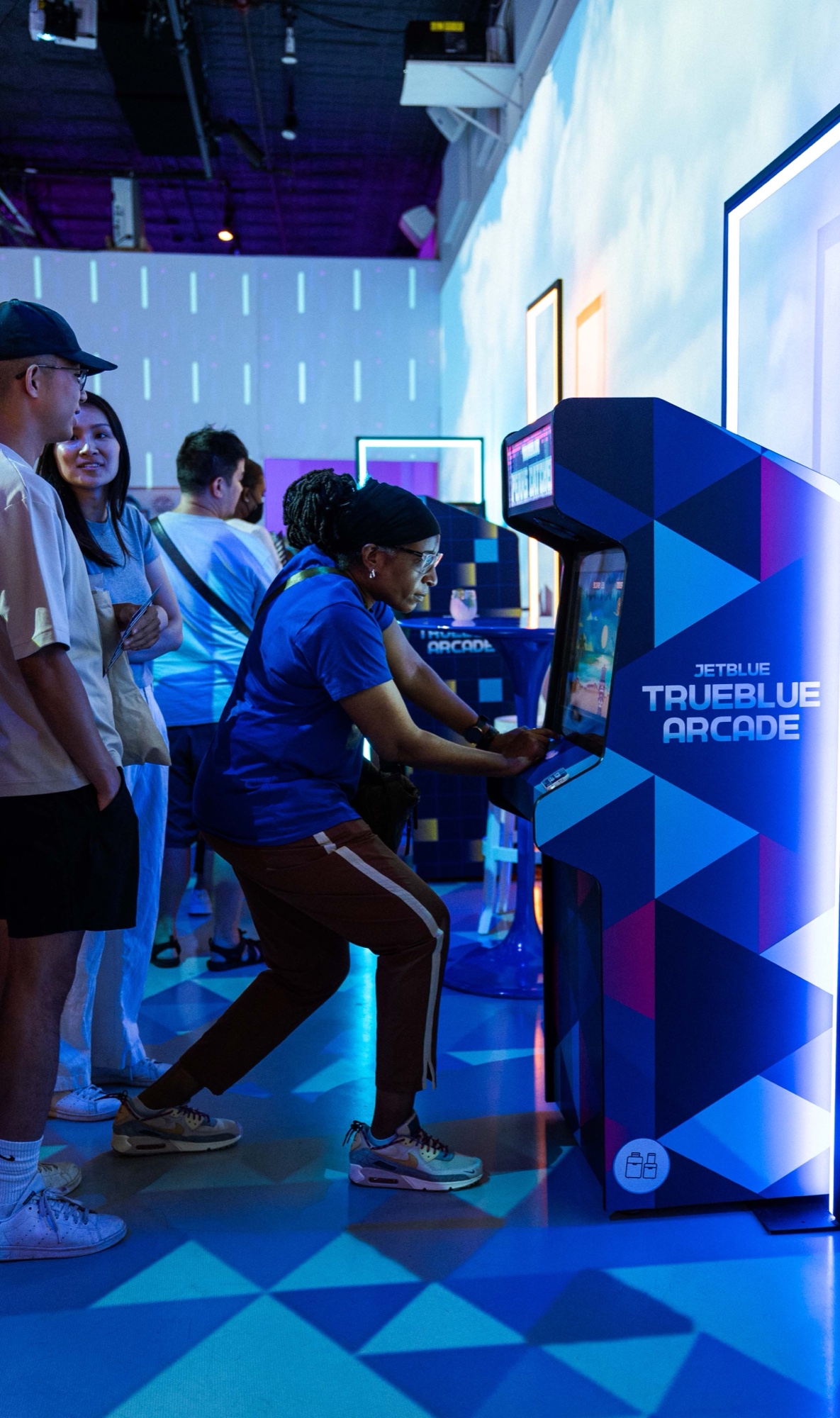 A woman in a blue shirt leans into a JetBlue TrueBlue arcade cabinet while friends watch in blue light