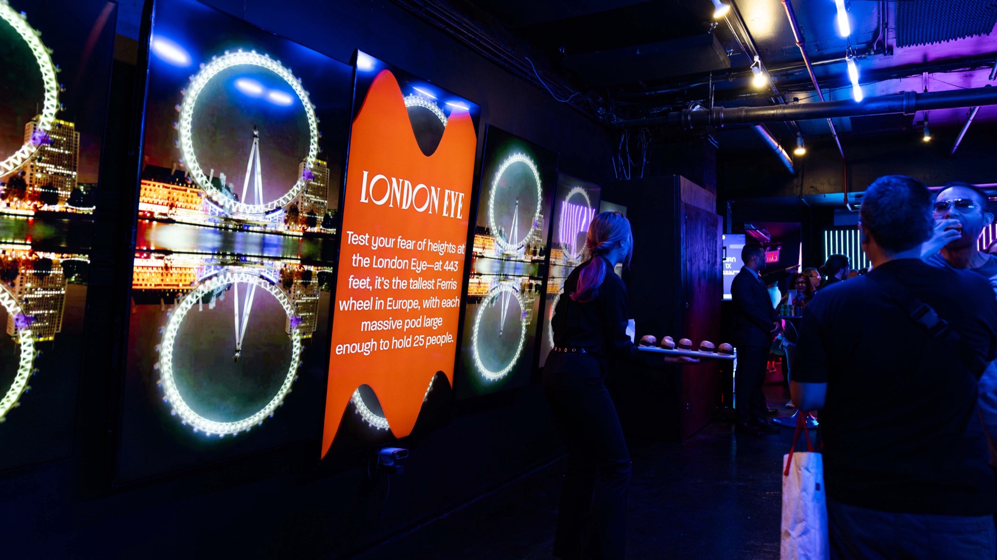 Guests beside a tall screen of the London Eye lit at night with an orange London Eye caption panel