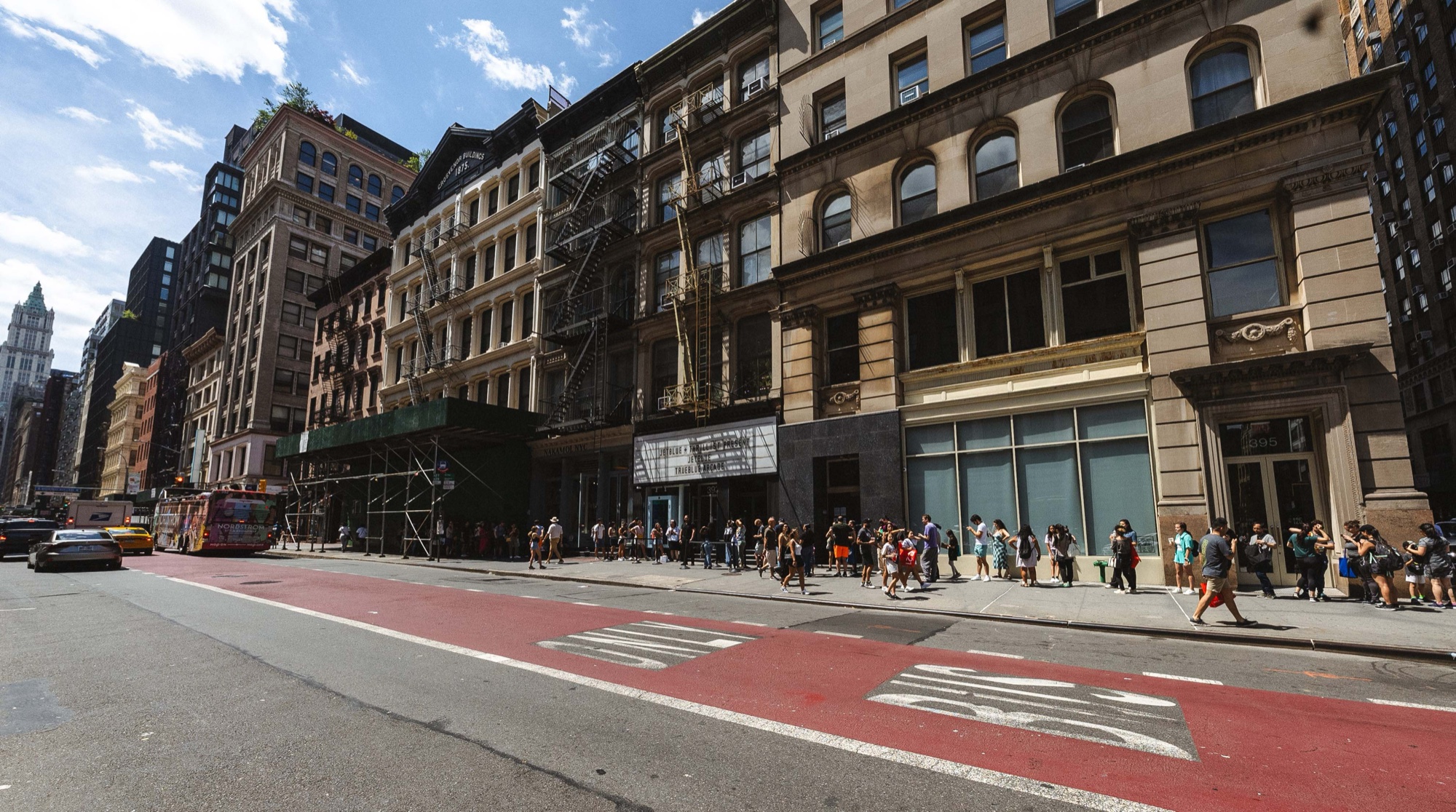 A queue of people stretches along a sunlit Manhattan sidewalk beneath old stone buildings and a red bus lane