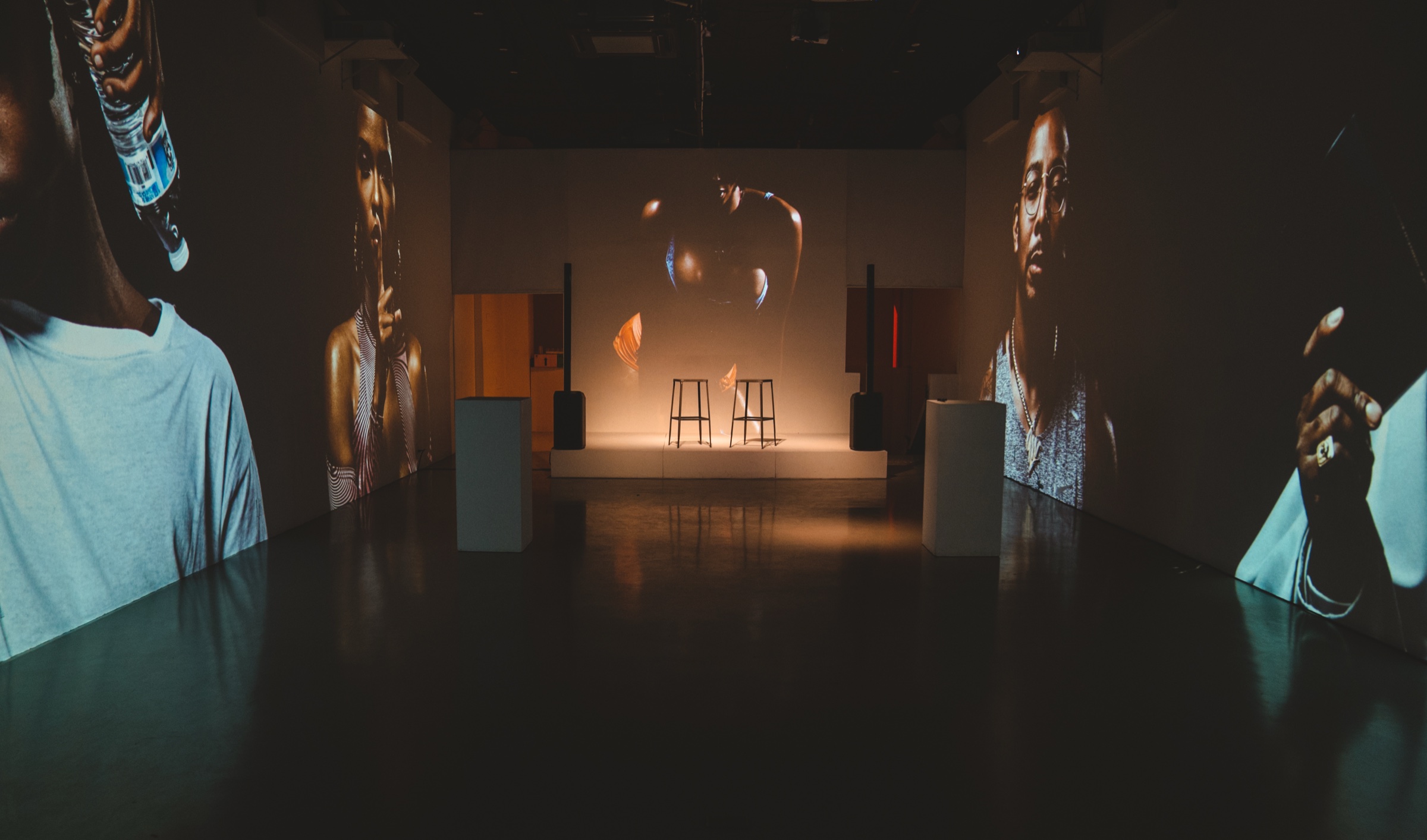 Dark room with two bar stools on a lit stage, large portraits projected on the side walls