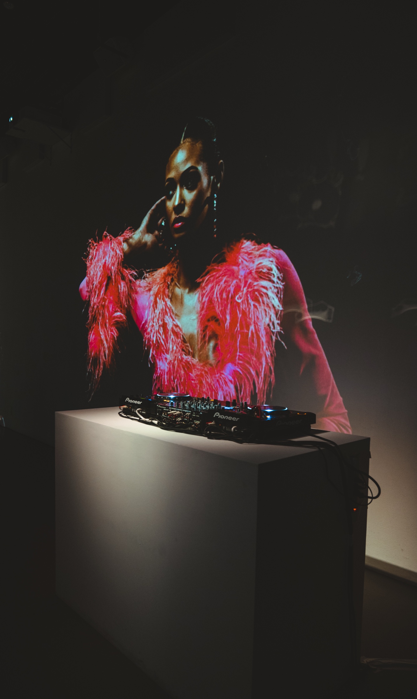Pioneer DJ decks on a white plinth before a projected portrait of a woman in a pink feathered coat
