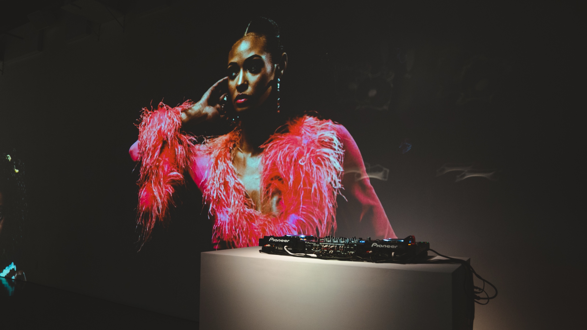 DJ decks on a white plinth in the dark beneath a projected portrait of a woman in pink feathers