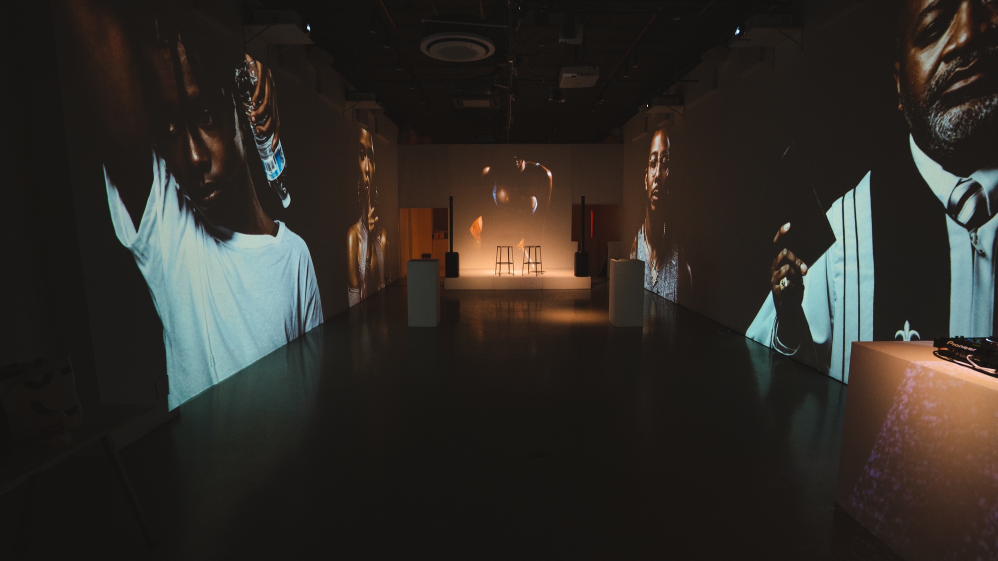 Dark hallway lined with large projected portraits, two stools and a lamp lit at the far end