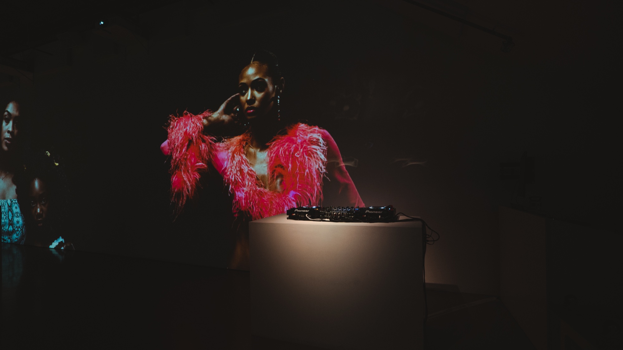 Projected portrait of a woman in a pink feathered jacket above a spotlit plinth with DJ decks