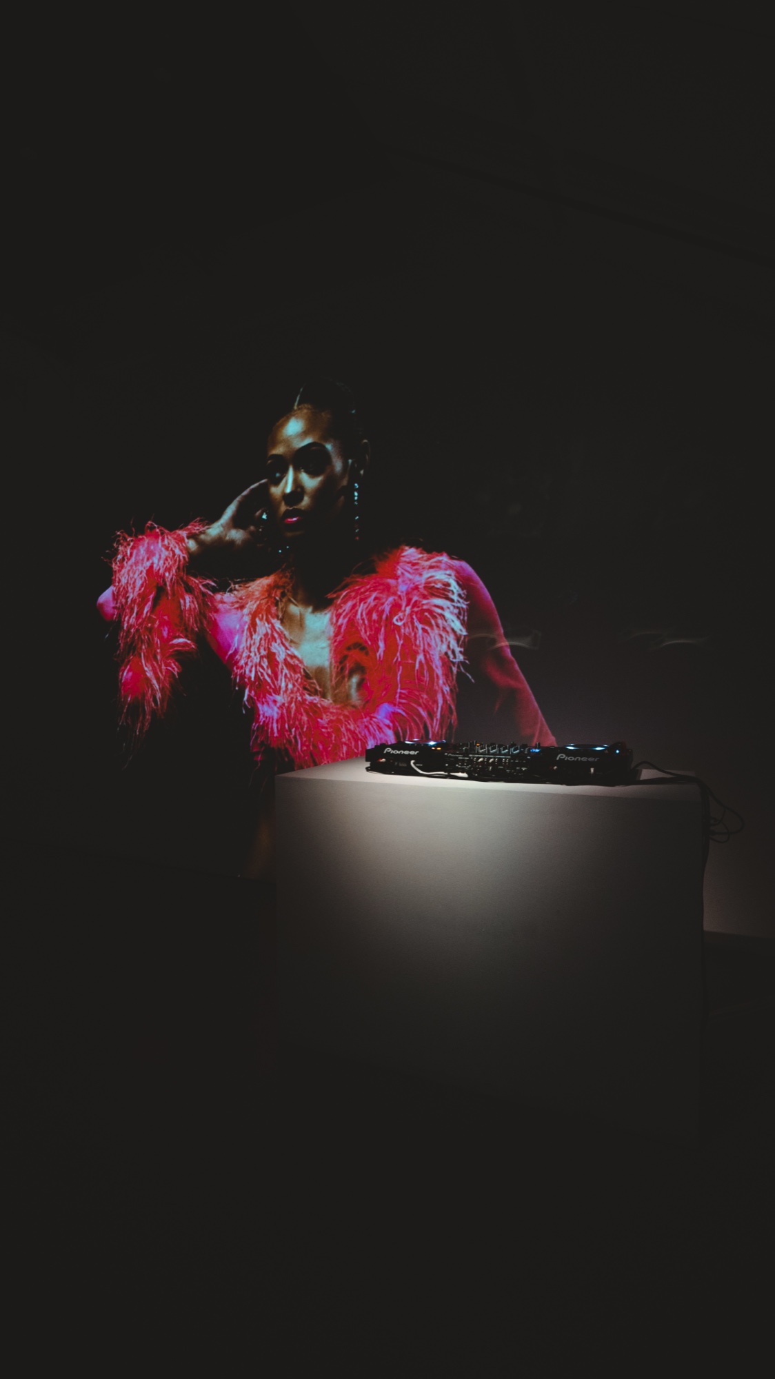 Woman in a feathered red jacket leans on a lit white plinth holding DJ decks in a dark room