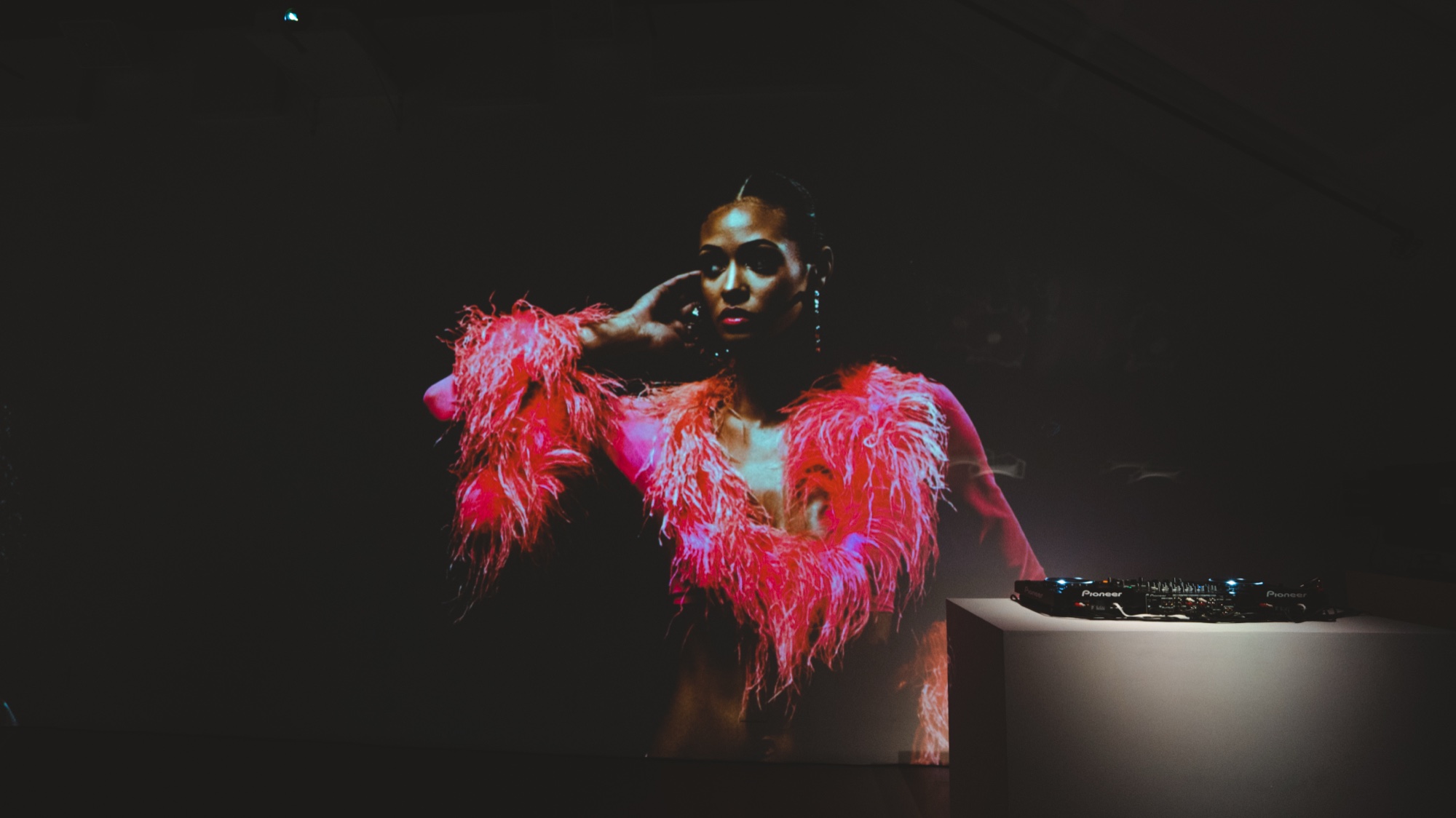 Projected portrait of a woman in a pink feathered coat glowing beside a DJ mixer on a white plinth