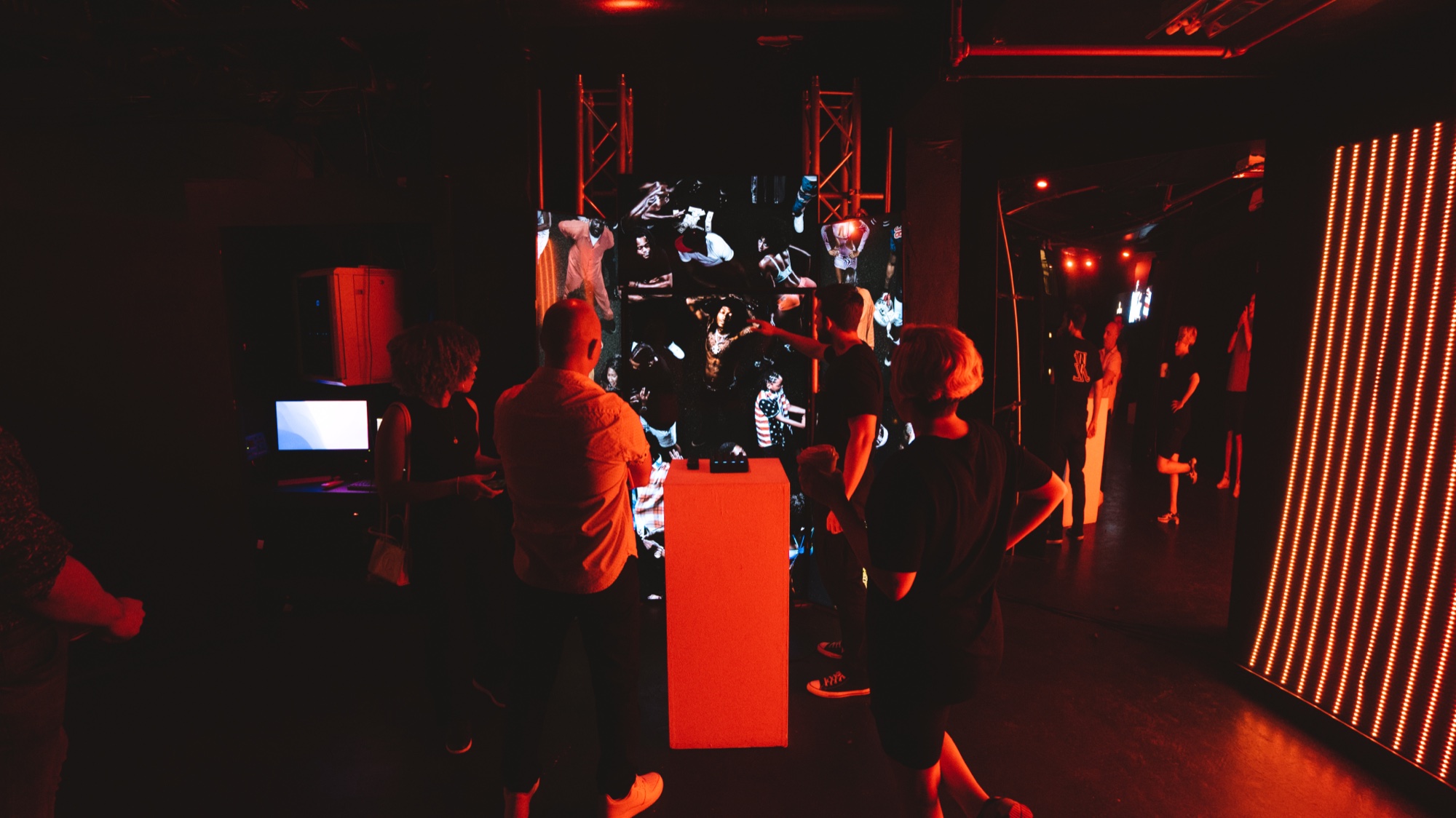 Guests in a dark room lit red watch a glowing screen beside a striped LED wall
