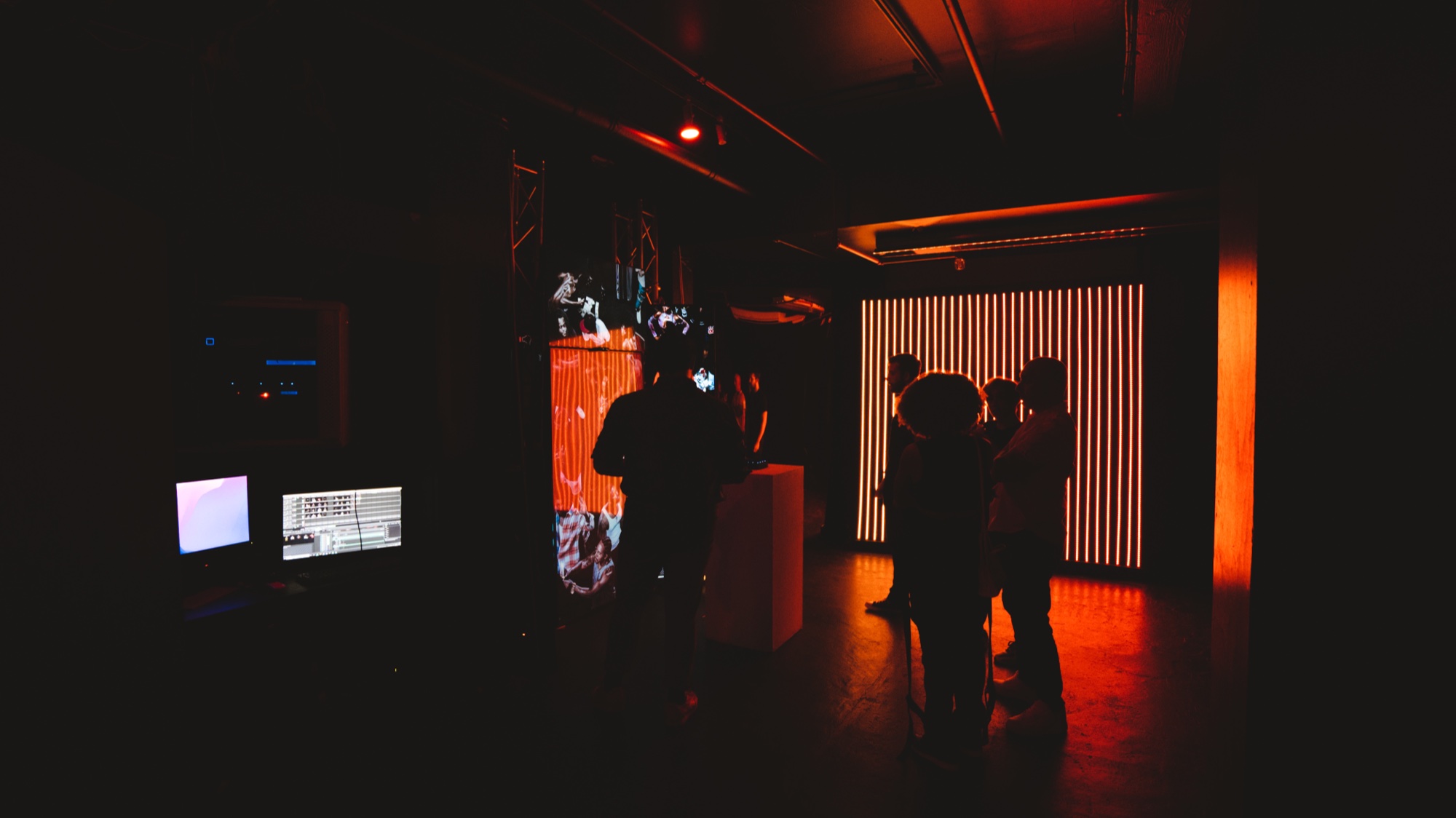 Silhouetted guests stand before an orange striped light wall in a dark room with a monitor glowing at left