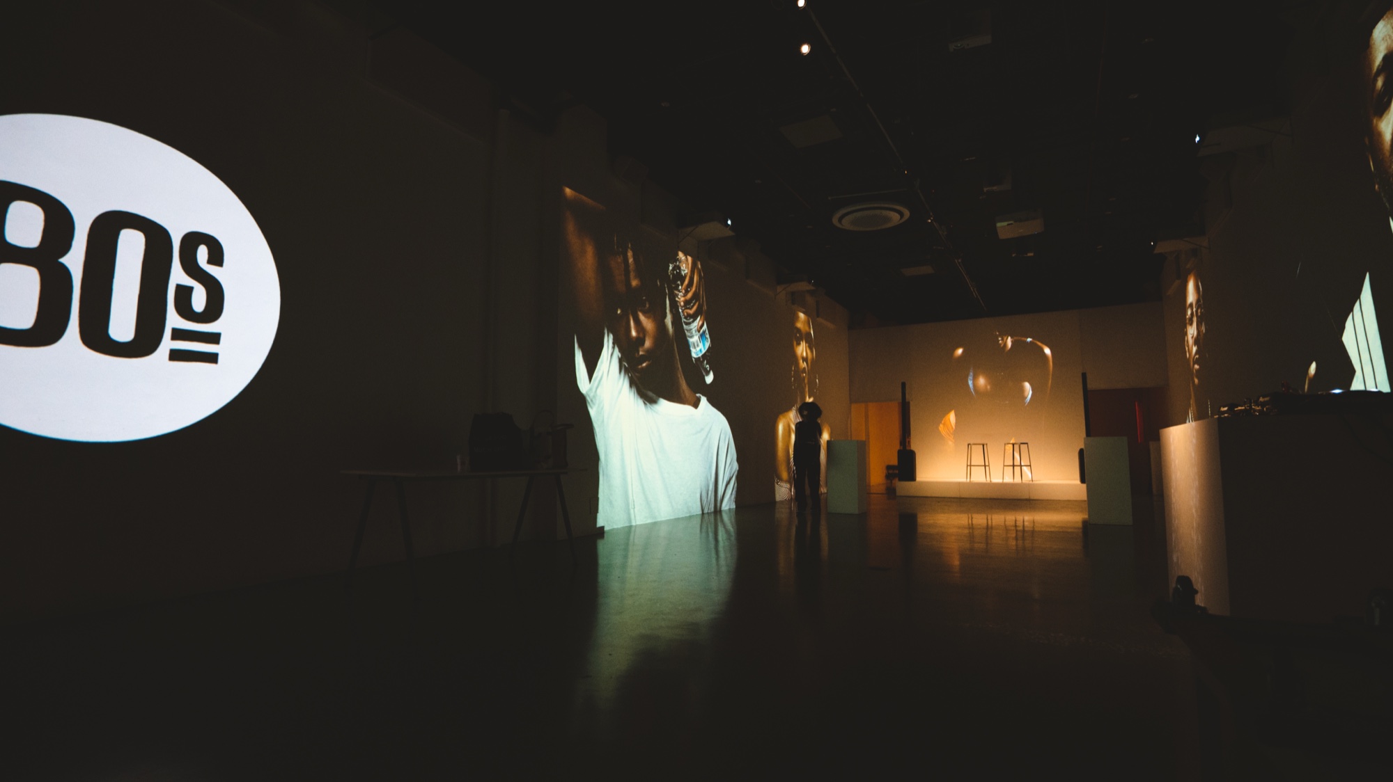 Dark gallery with an 80s logo on a white circle and tall projected figures along the walls