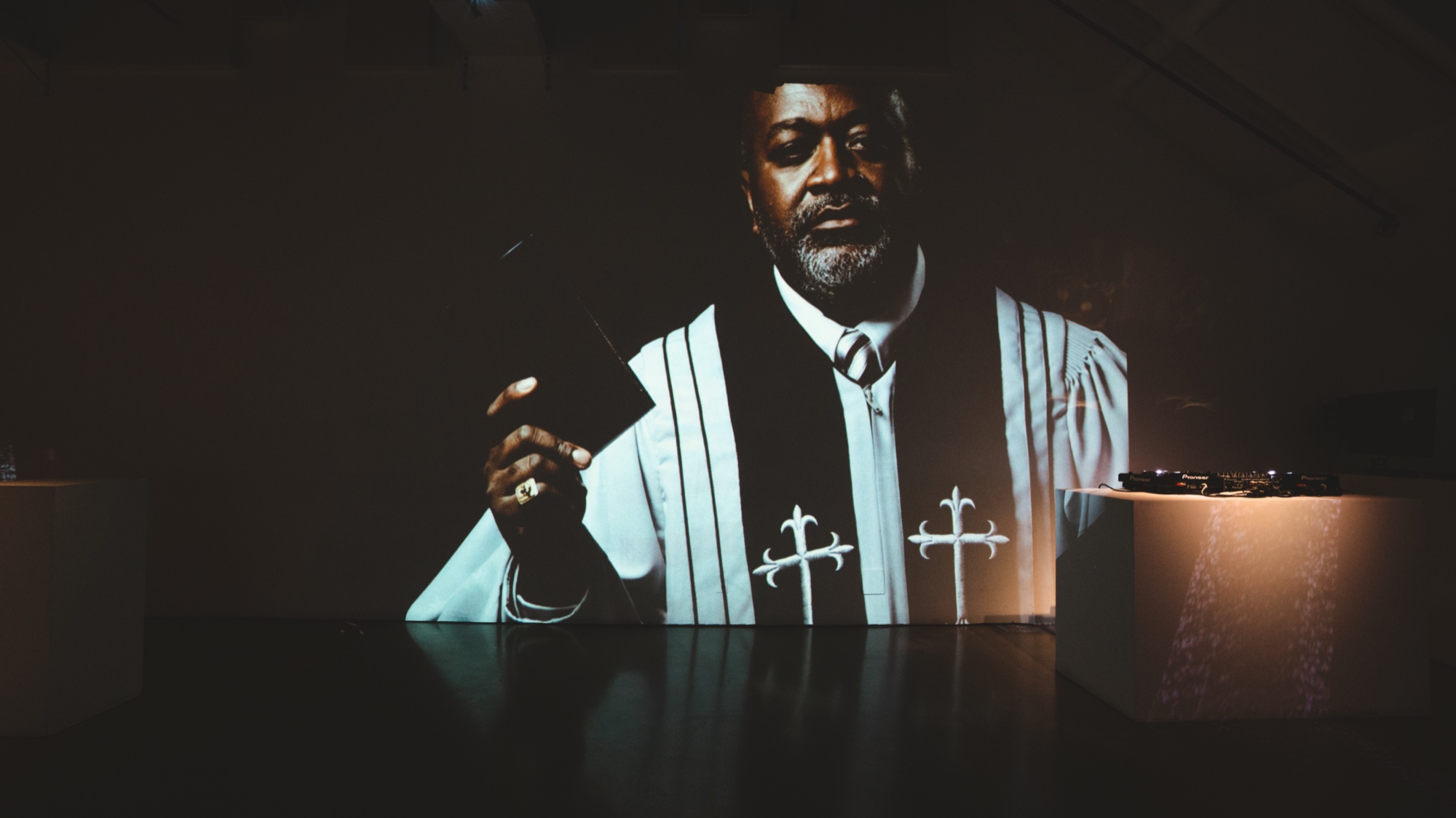 Projection of a man in white choir robes with silver crosses raising a phone in a dark room
