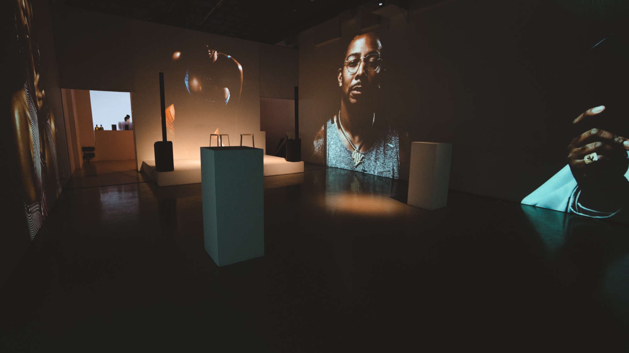 Dark room with plinths, a lit stage with two stools and a projected portrait of a man in glasses