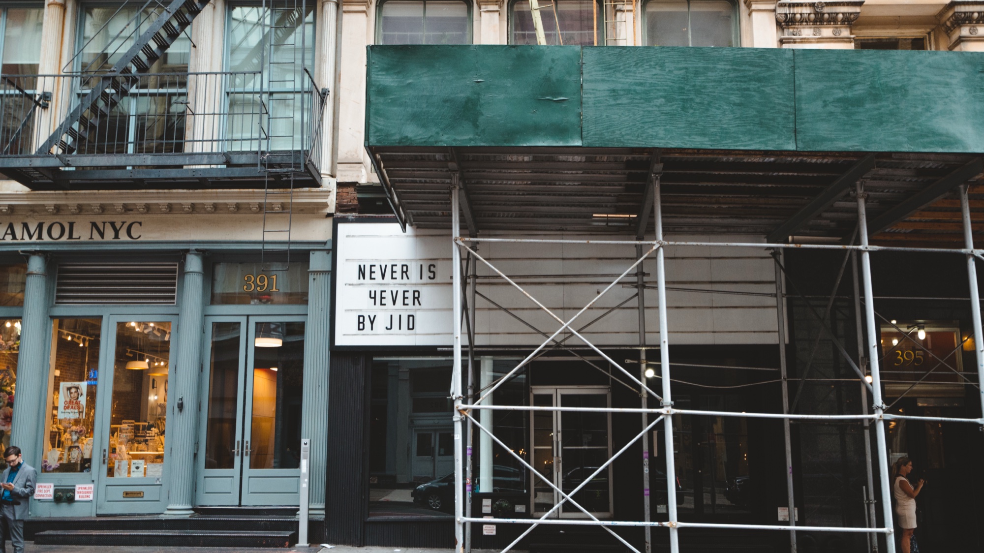 Marquee sign reading Never Is 4ever By JID under green scaffolding beside a cast iron storefront