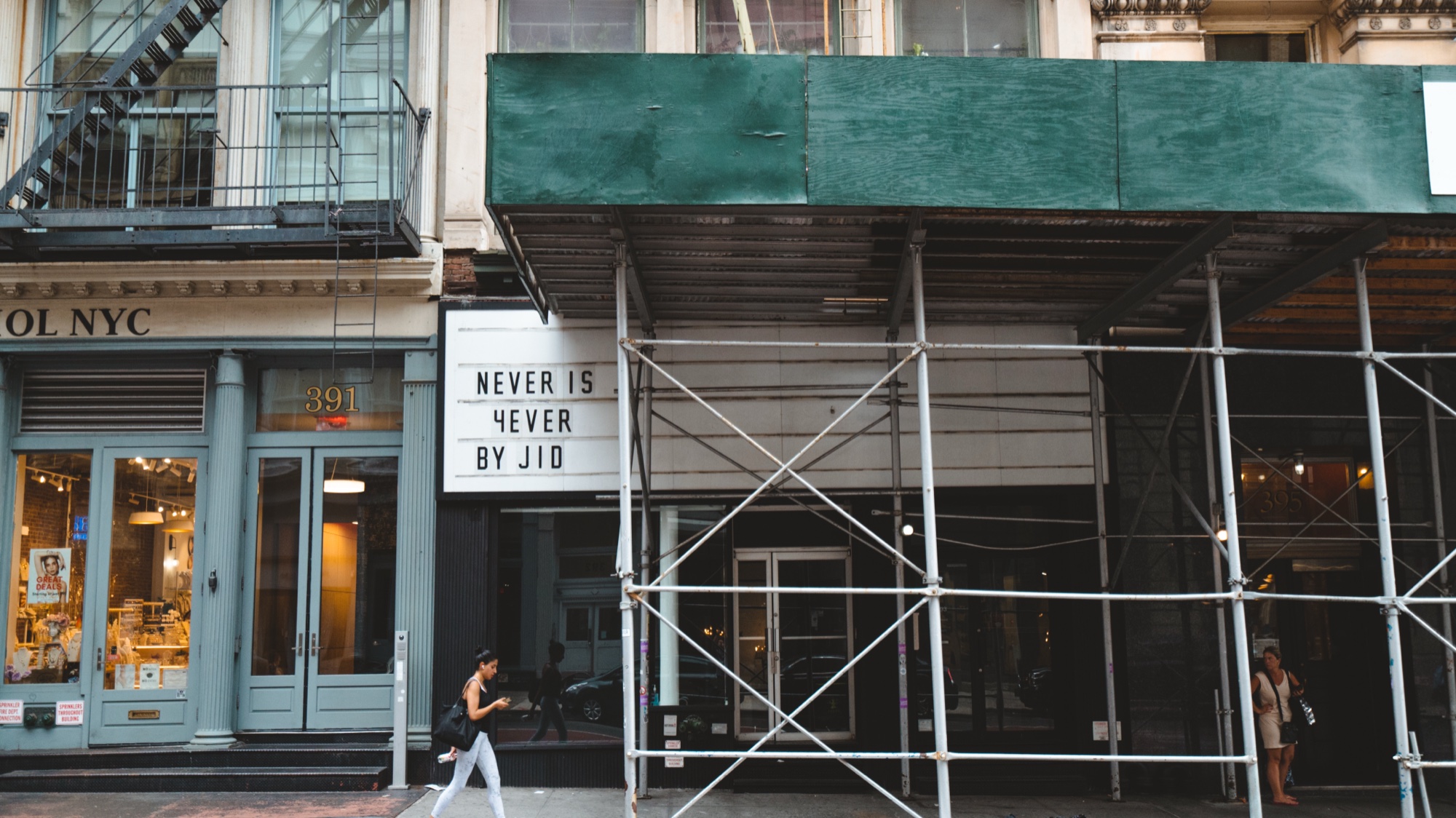 Woman walks past a marquee reading Never Is 4ever By JID beneath green scaffolding on a city block