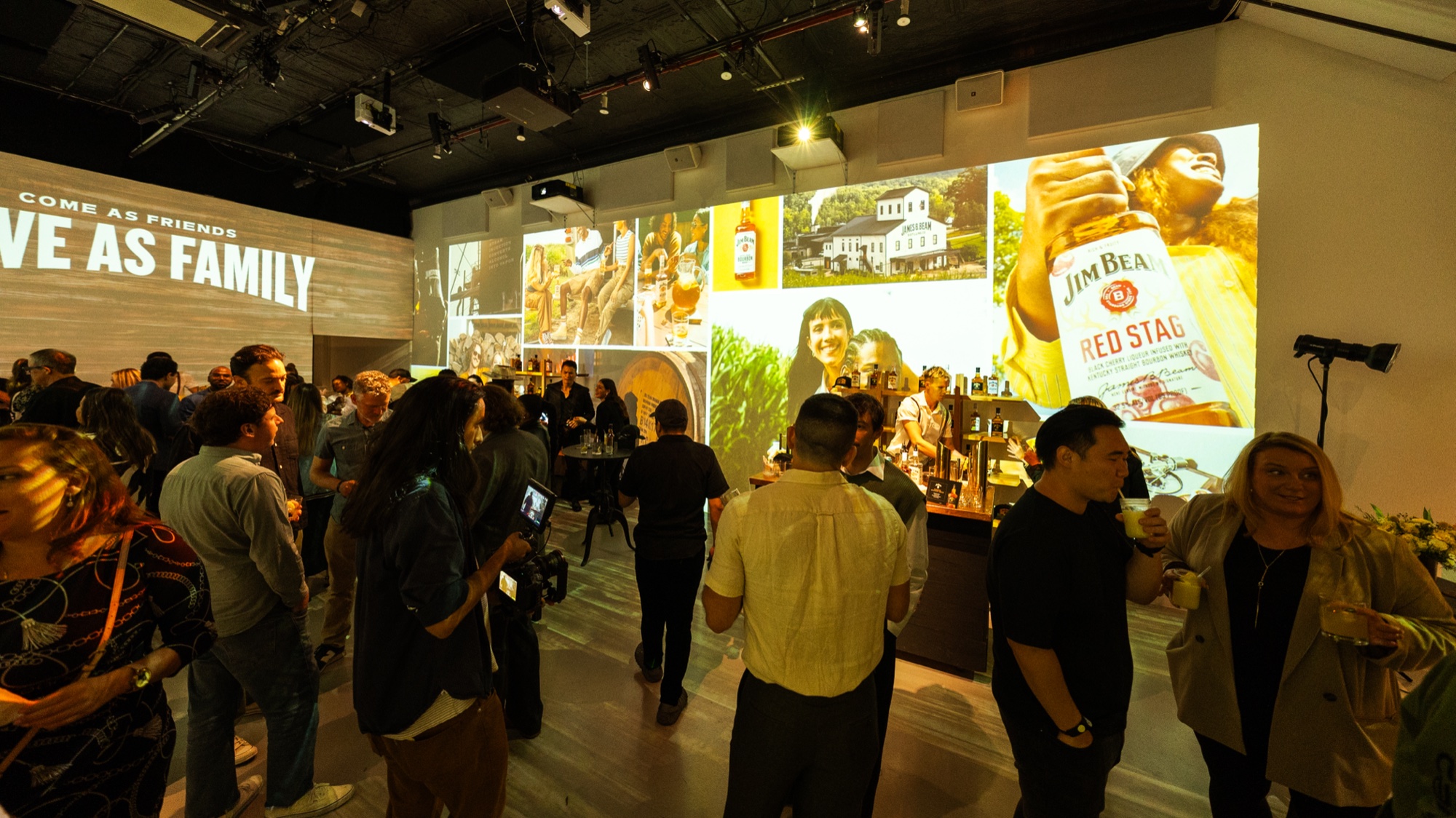 Crowd mingles beneath a wall projection reading Leave As Family beside a Jim Beam Red Stag bottle