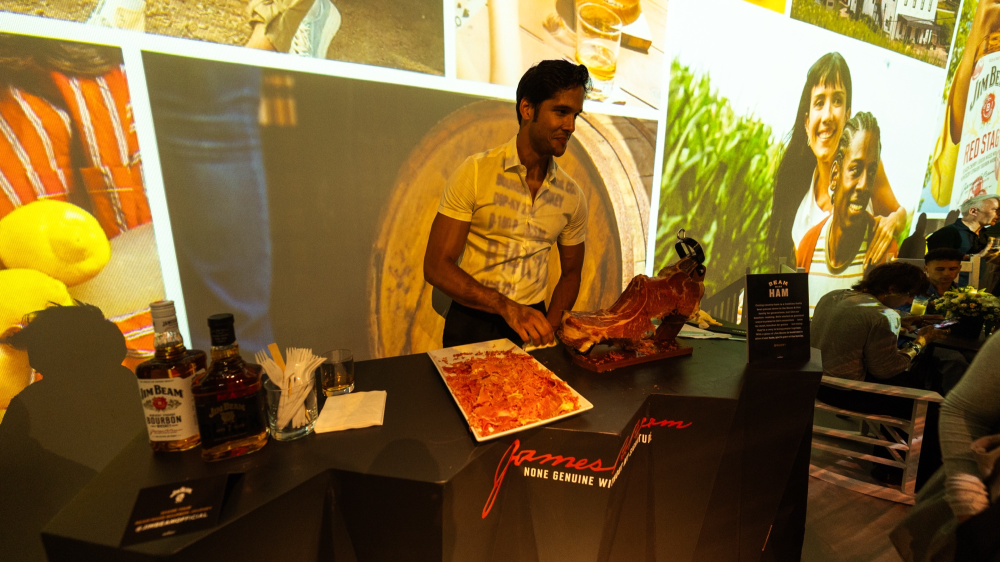 Man behind a black bar slices cured ham next to Jim Beam bottles and lemons under projected imagery