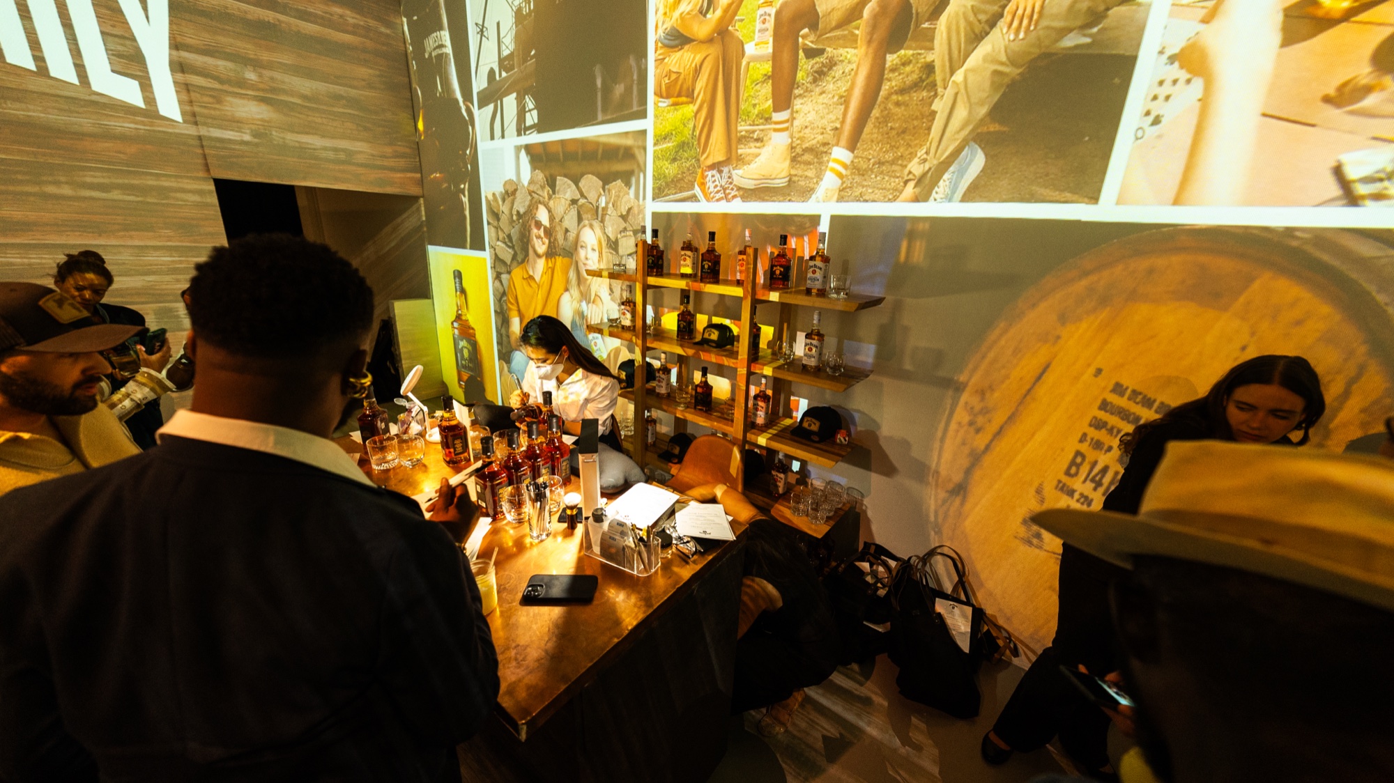 Bartender in a white shirt works a wooden bar beside a whiskey barrel under projected photos of denim and boots