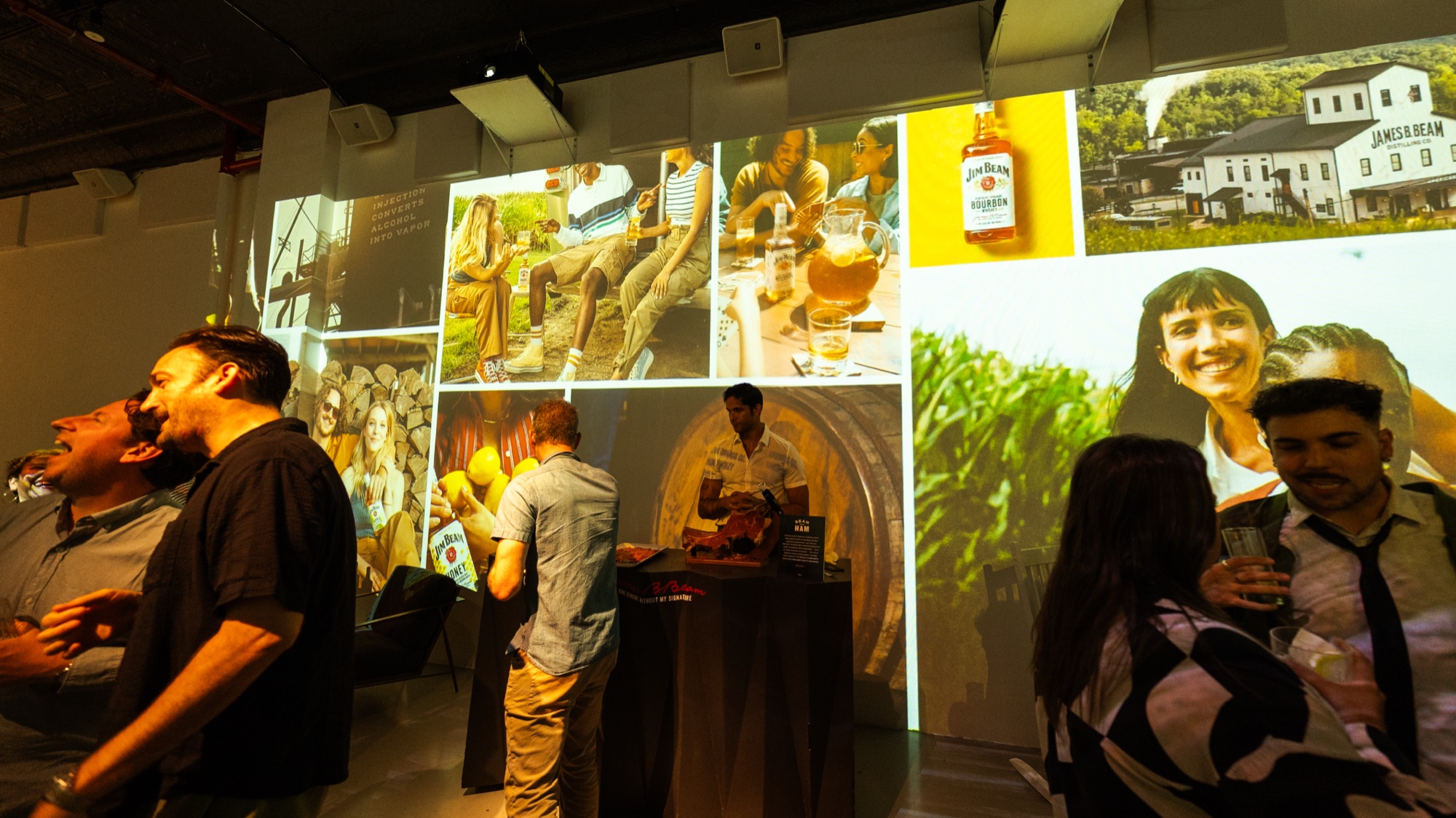 Guests drink and talk before a wall of projected photographs and a Jim Beam bottle