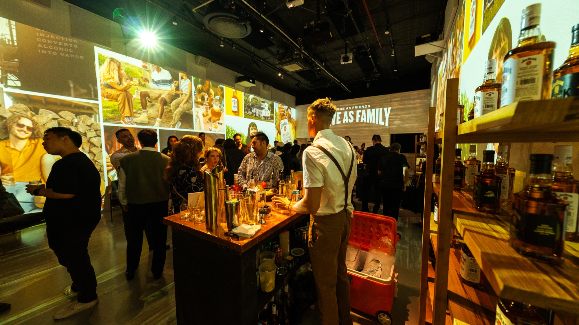 Bartender in suspenders works a wooden bar as guests mill under a Leave As Family projection