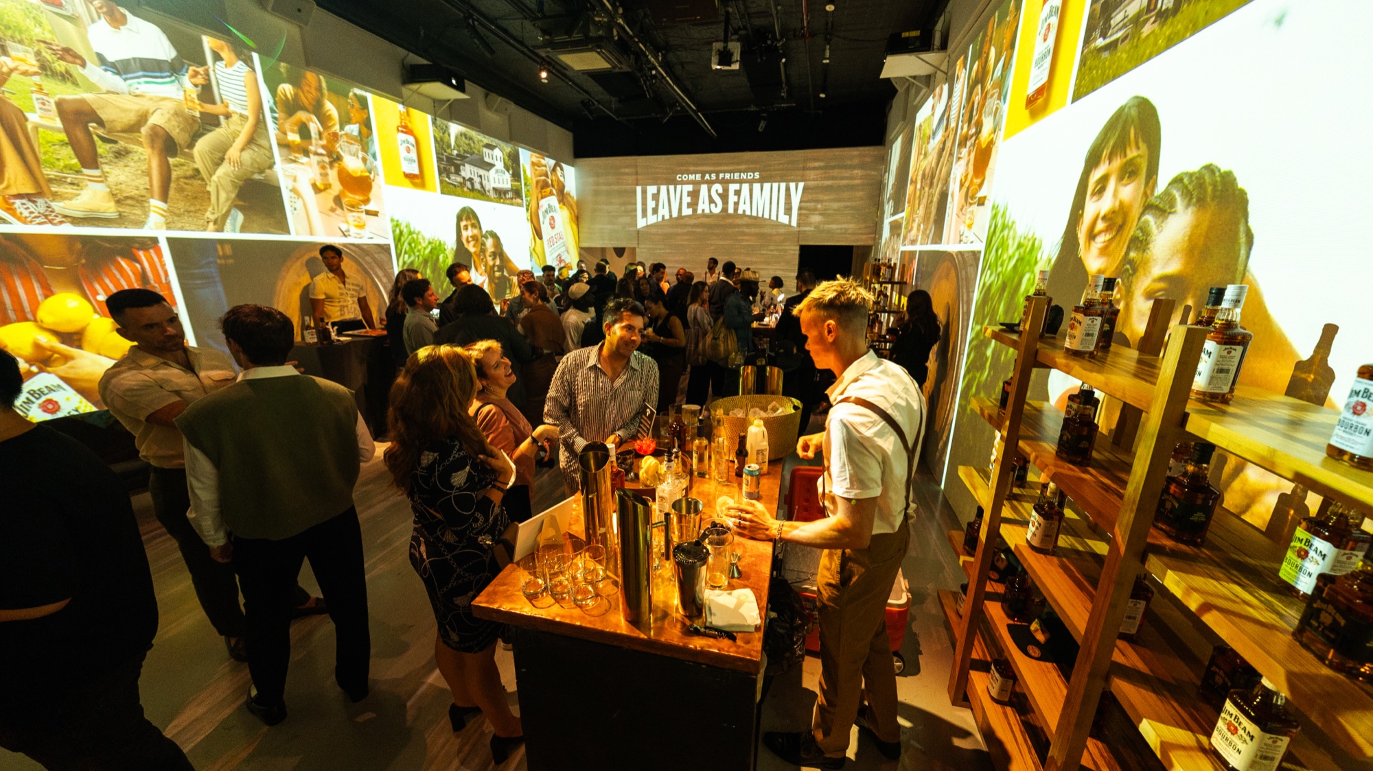 Bartender in a white shirt serves drinks at a wooden bar beneath projections reading Leave As Family