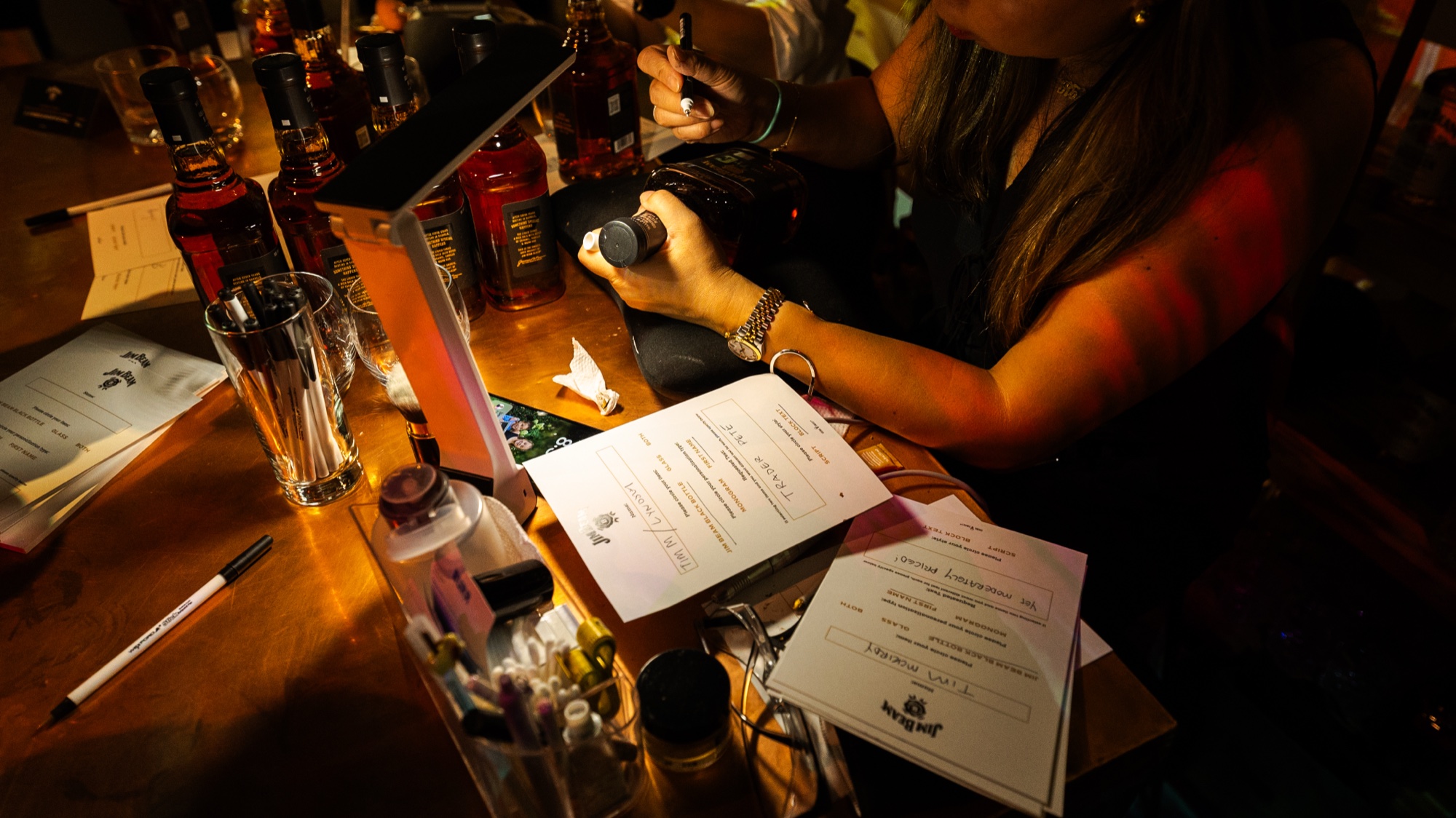 Hands and tasting note cards on a warmly lit wooden bar lined with whiskey bottles and a candle