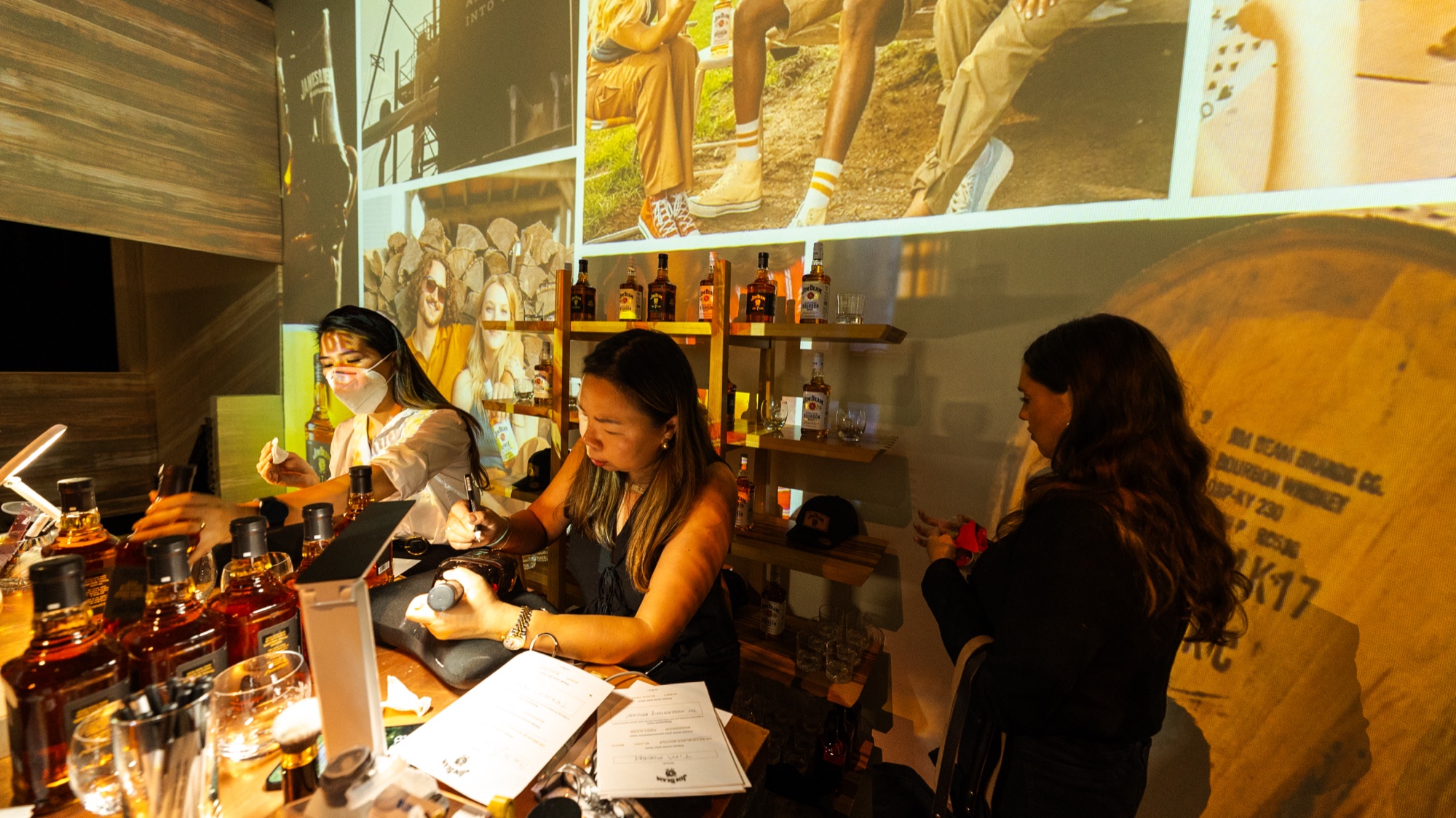 Two women work behind a bar of whiskey bottles next to a large barrel, projected photos overhead