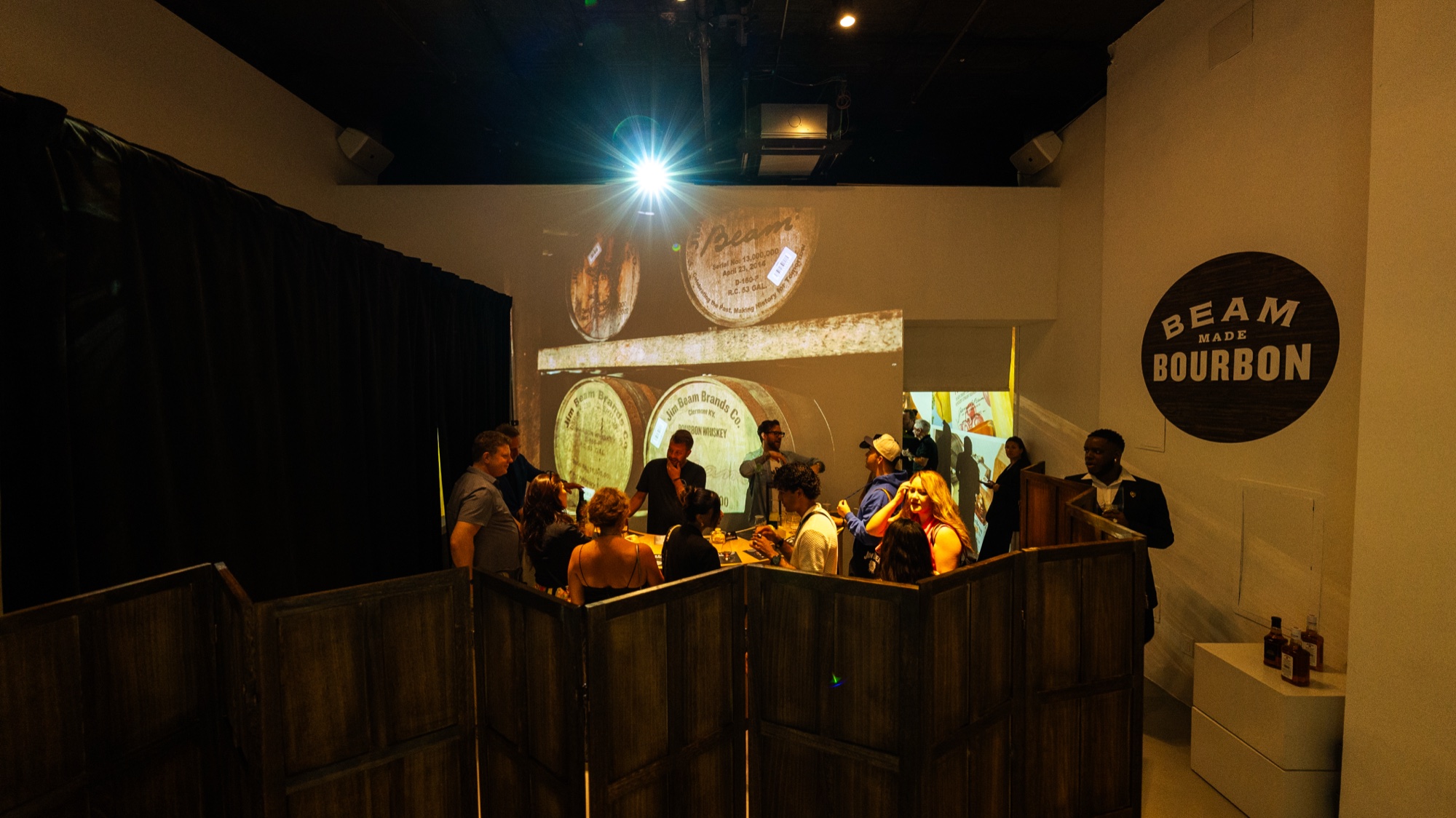 Guests seated around a wooden bar under projected barrel ends and a round Beam Made Bourbon sign