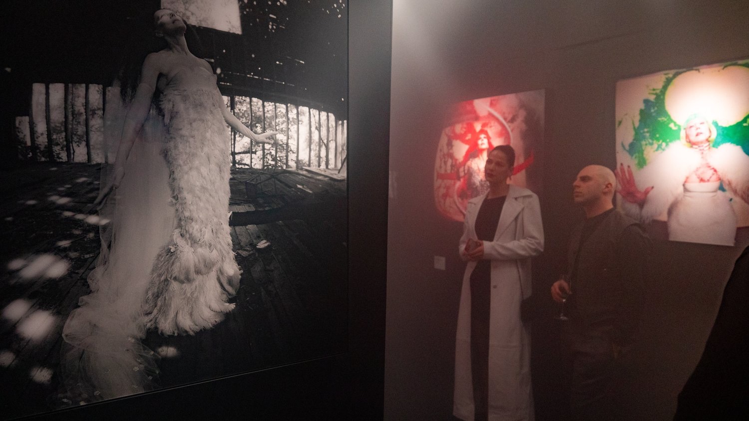 Portrait featuring a black and white feathered gown beside guests viewing colorful works at the exhibition