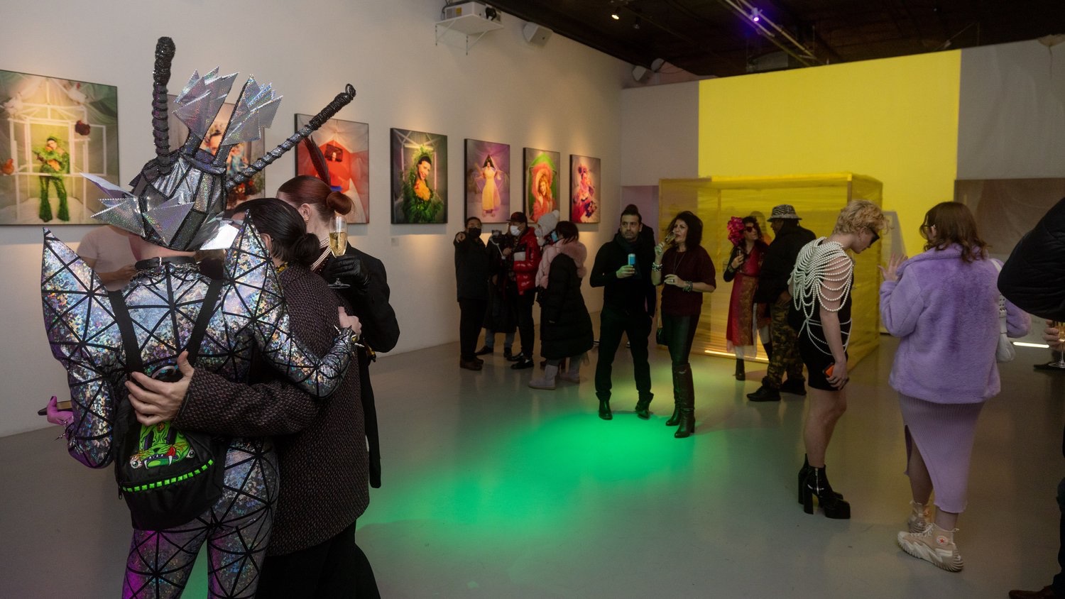 Guest in a sparkling spiked costume embraces a friend near pastel portraits and a yellow wall