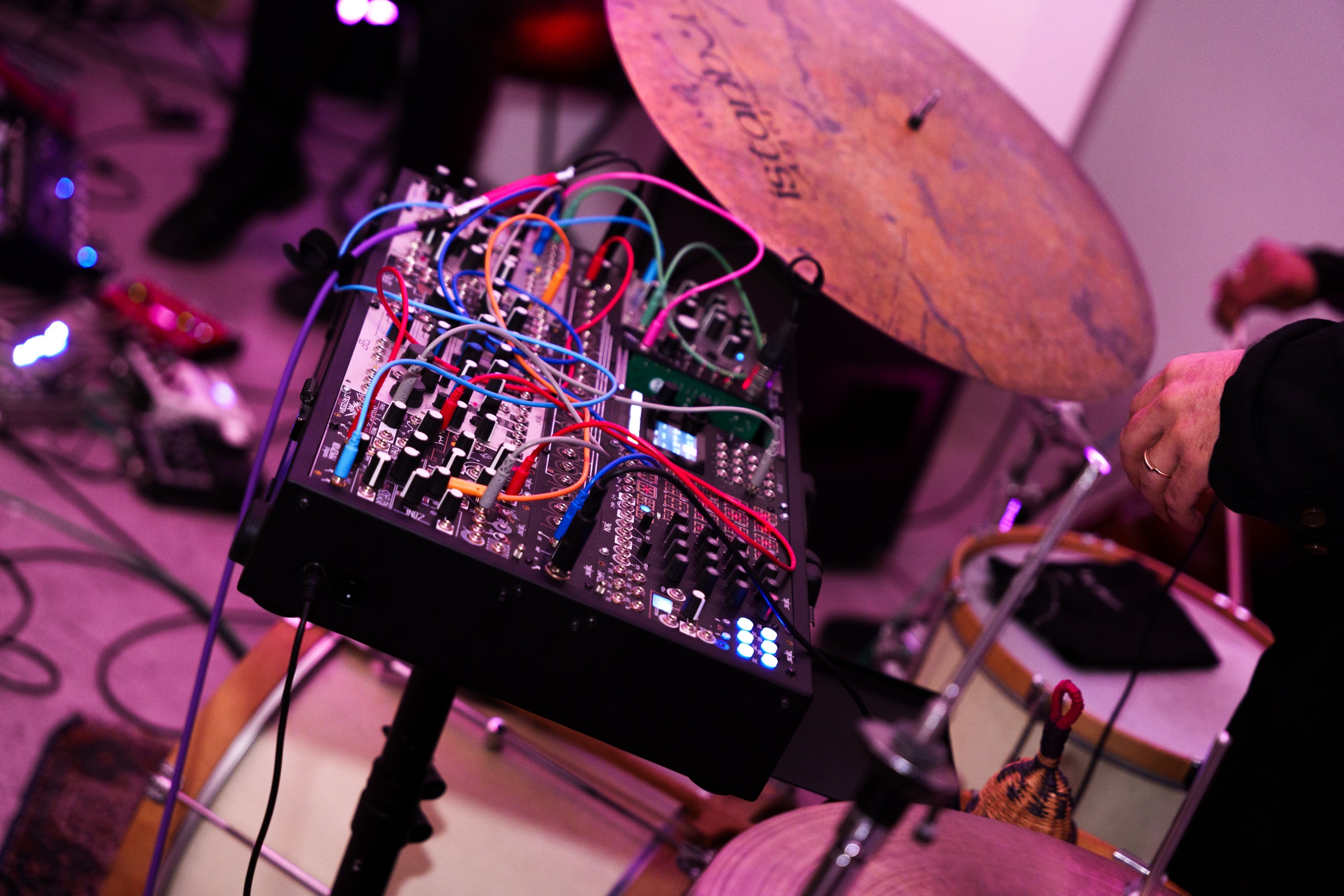 A modular synthesizer tangled with colourful patch cables sits beside a cymbal and drum kit under pink light