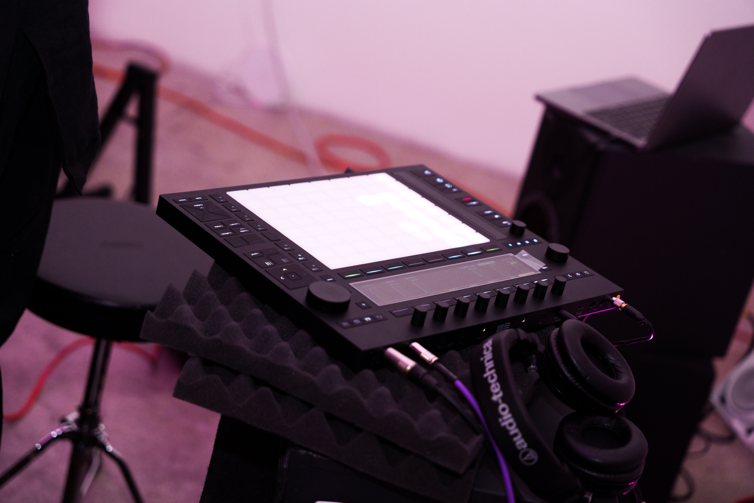 Black sampler pad and headphones resting on foam pads beside a stand in a pink lit studio