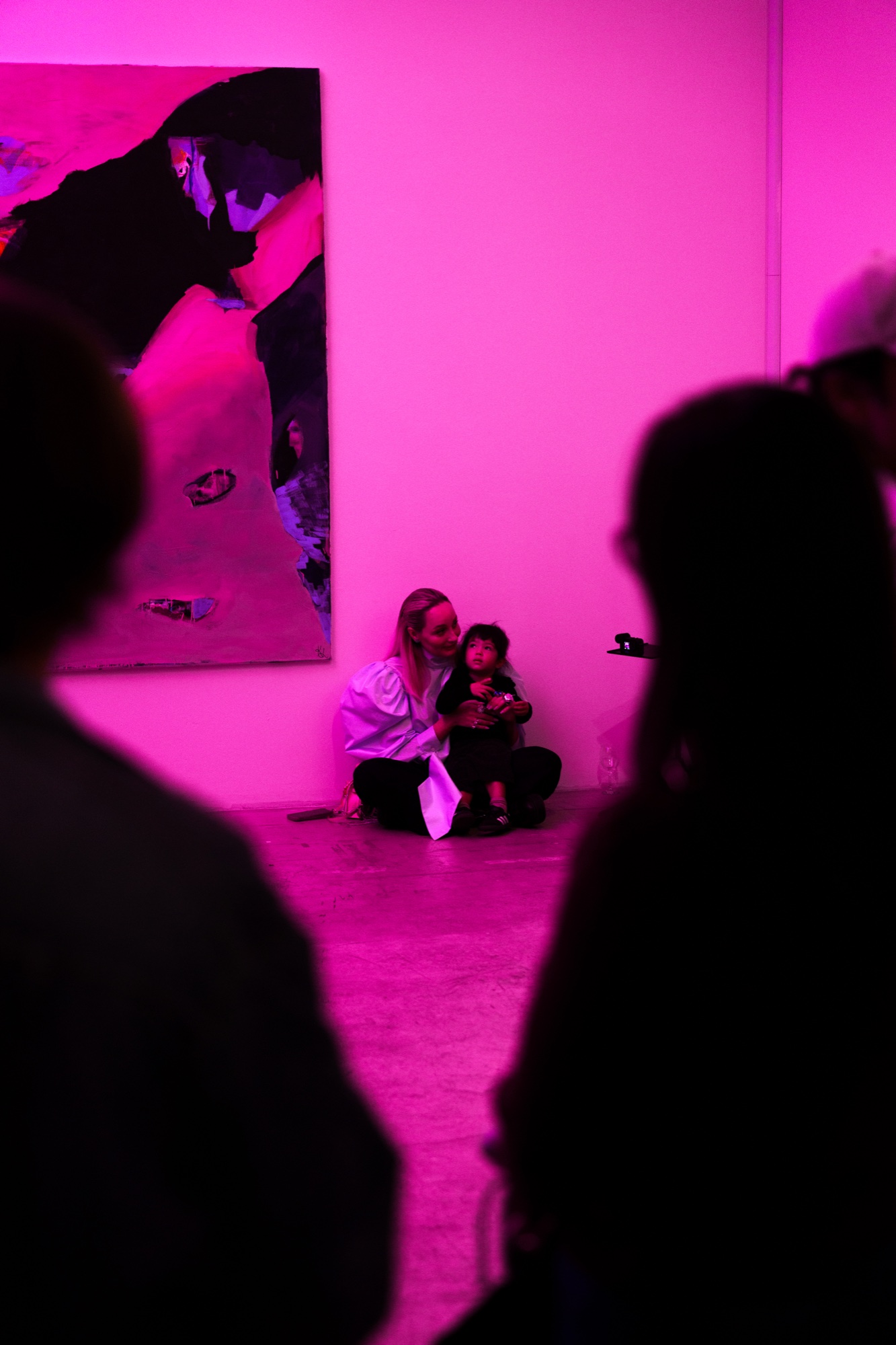 A woman sits on the floor with a small child beneath a large abstract painting in a pink lit room