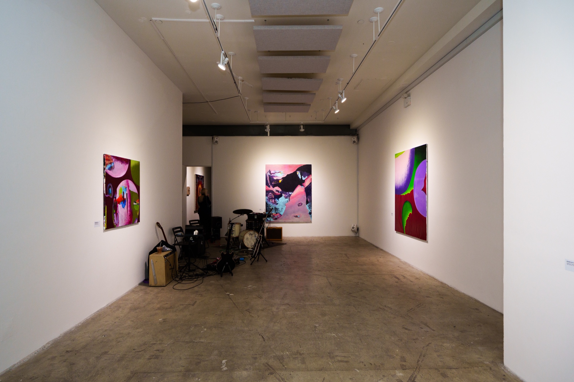 A white gallery with three colourful abstract paintings on the walls and a drum kit at the far end