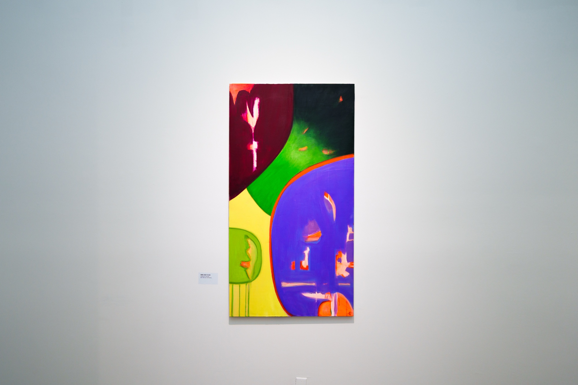A tall abstract painting in purple, green, yellow and dark red hangs alone on a pale gallery wall