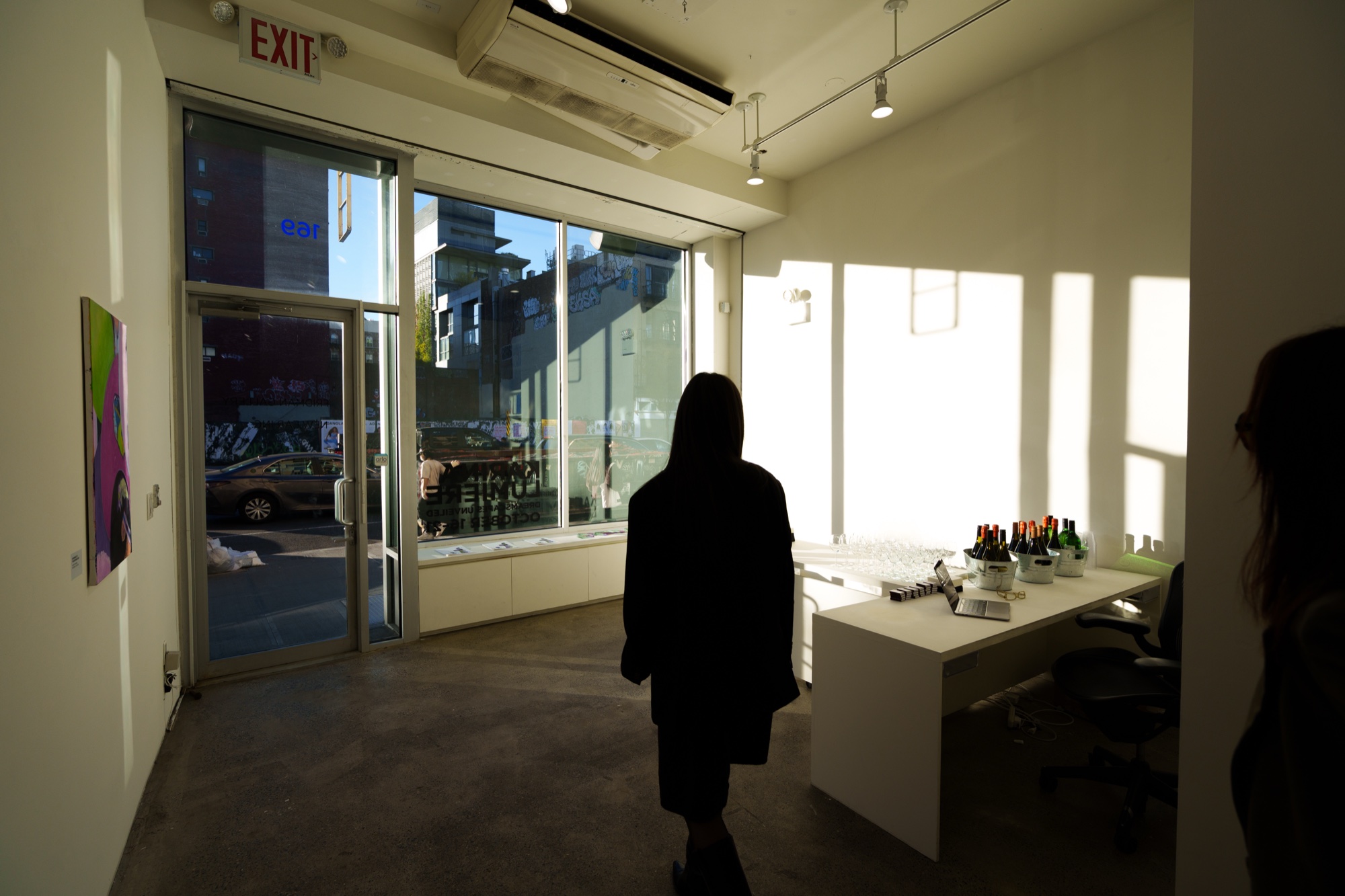 A silhouetted visitor walks toward a sunlit gallery storefront window with bottles set out on a white desk