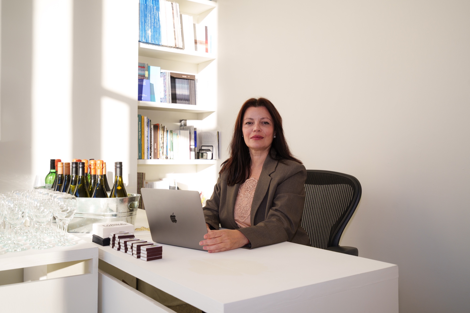 A woman in a brown blazer sits at a white desk with an open laptop, wine bottles and bookshelves behind