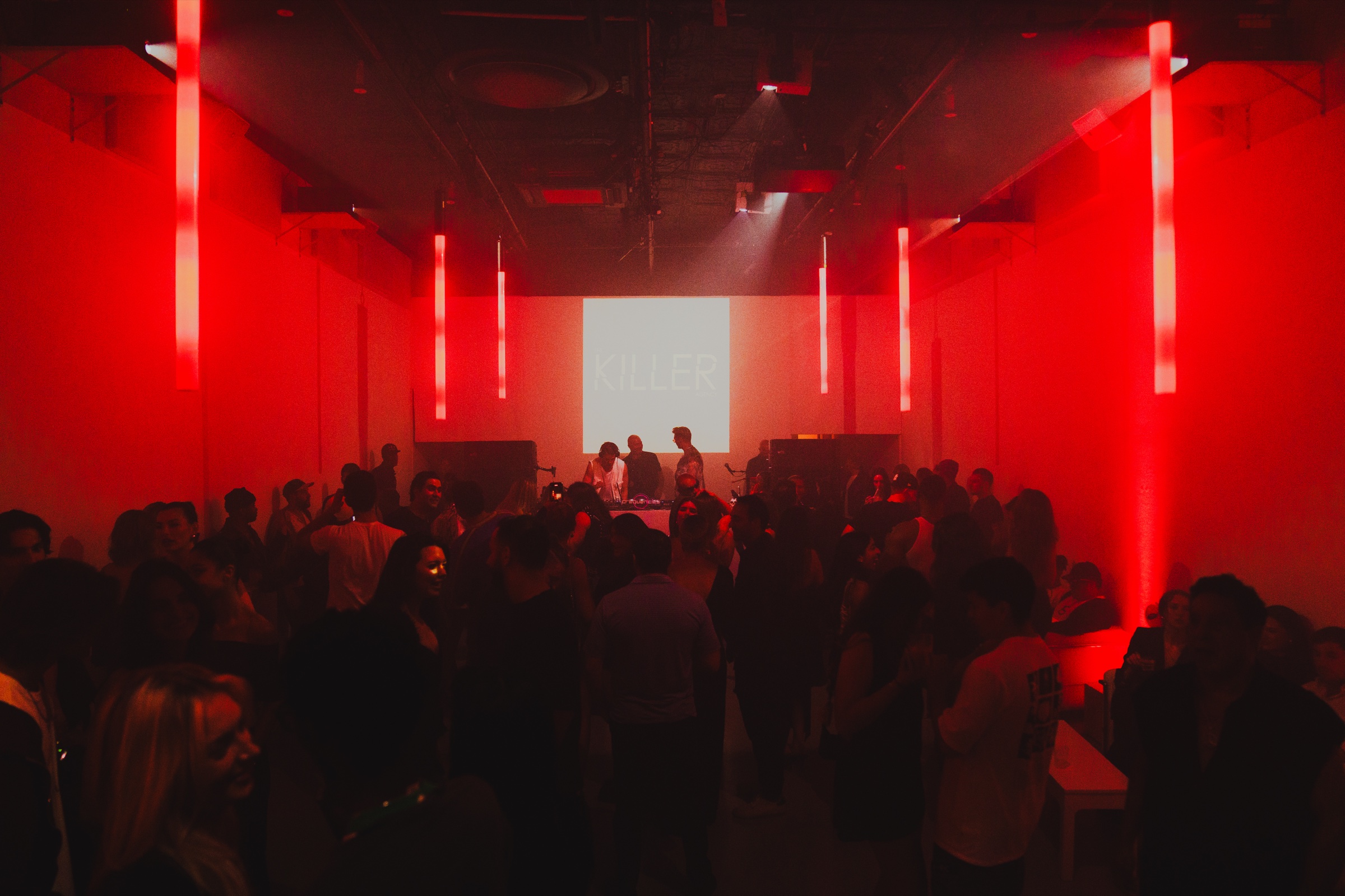 A crowd packs a red lit room with vertical red light tubes and two DJs beneath a bright projection screen