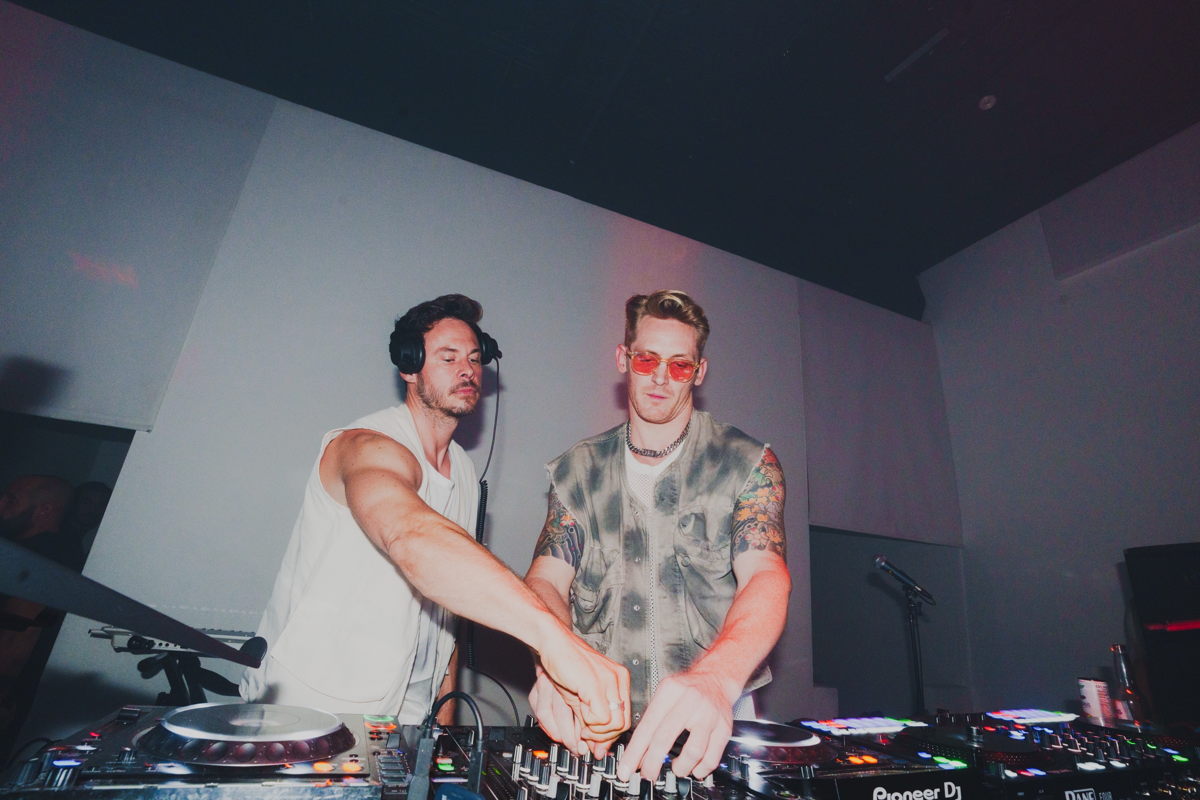 Two DJs in vests and headphones operate Pioneer decks with colorful glowing buttons at The Killer Agency event