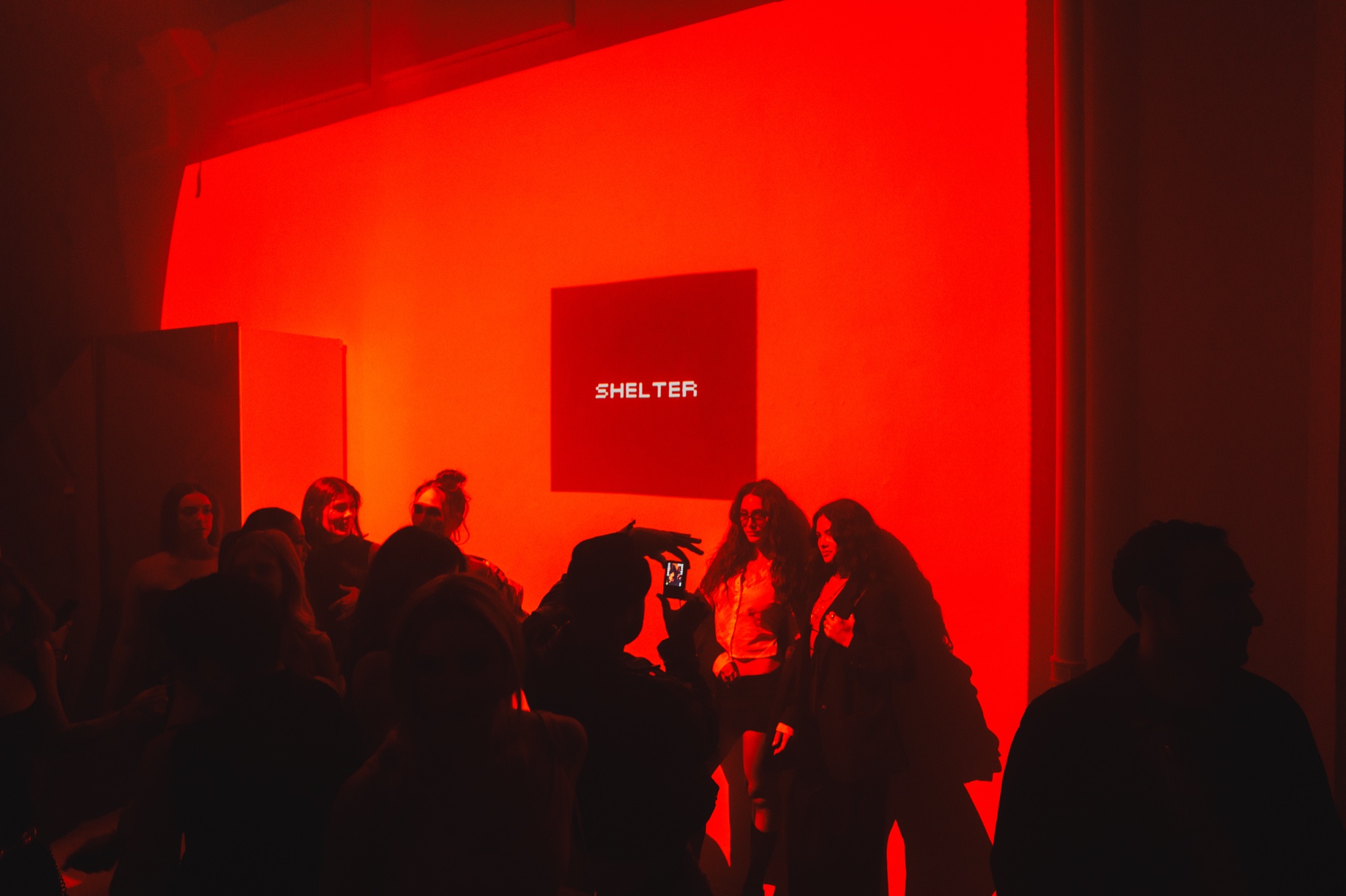 Guests in silhouette against a red lit wall with a small square screen reading Shelter
