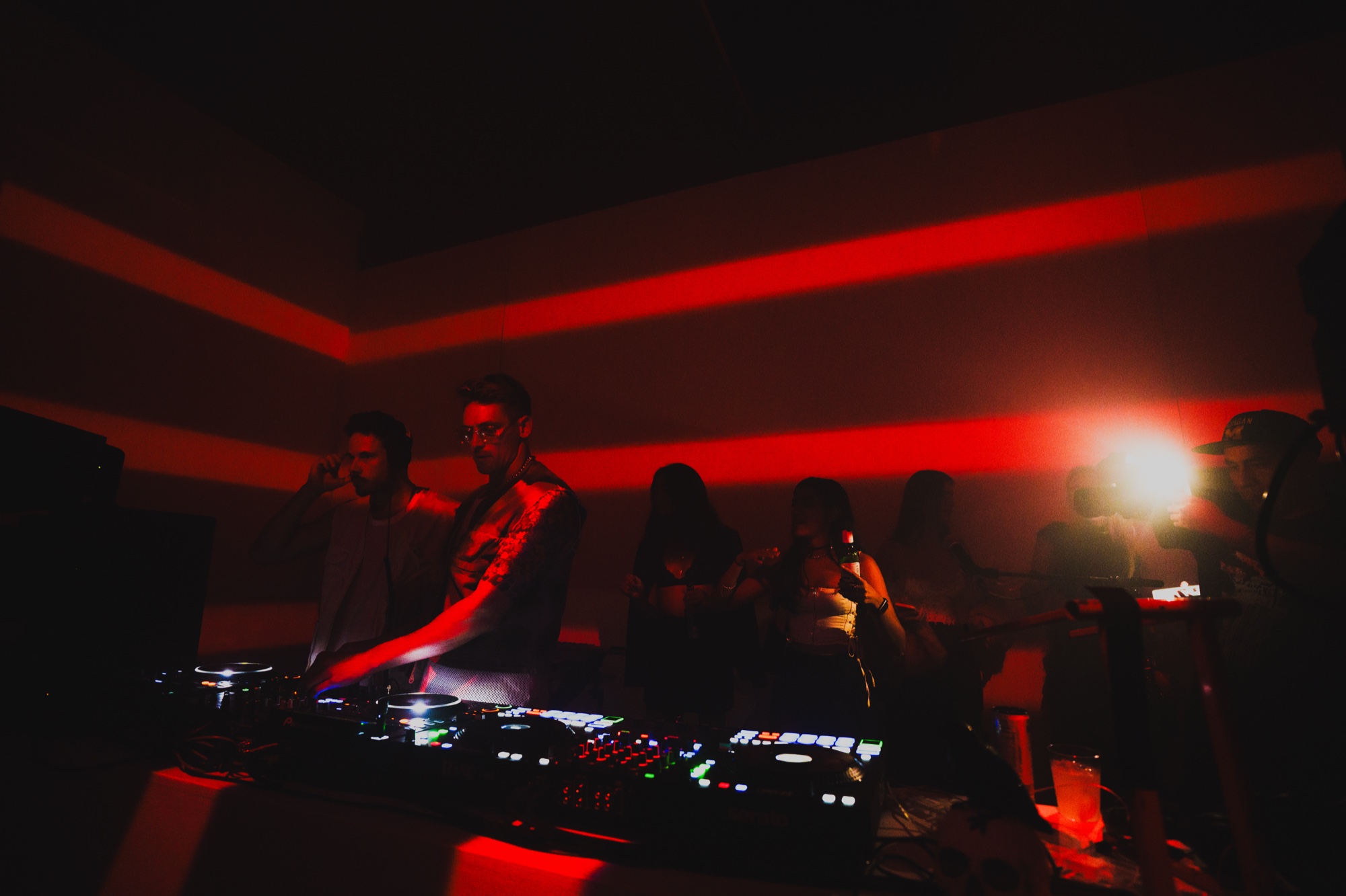 Two DJs at a mixer in a dark room banded with red light, silhouetted guests behind