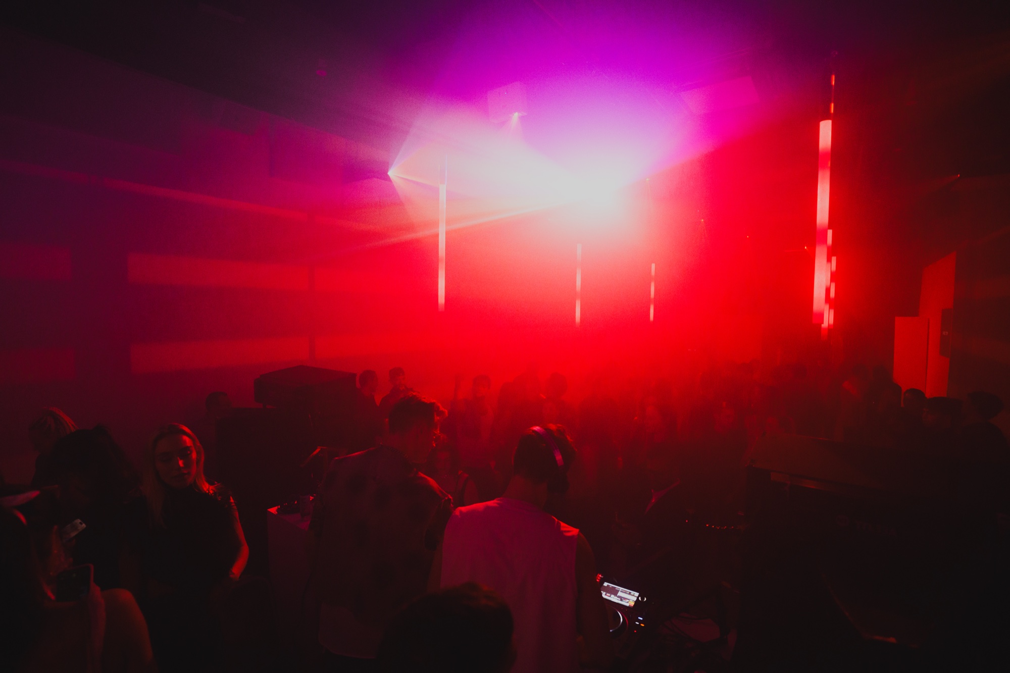 A crowd in silhouette faces a bright white light through red and magenta haze in a dark club