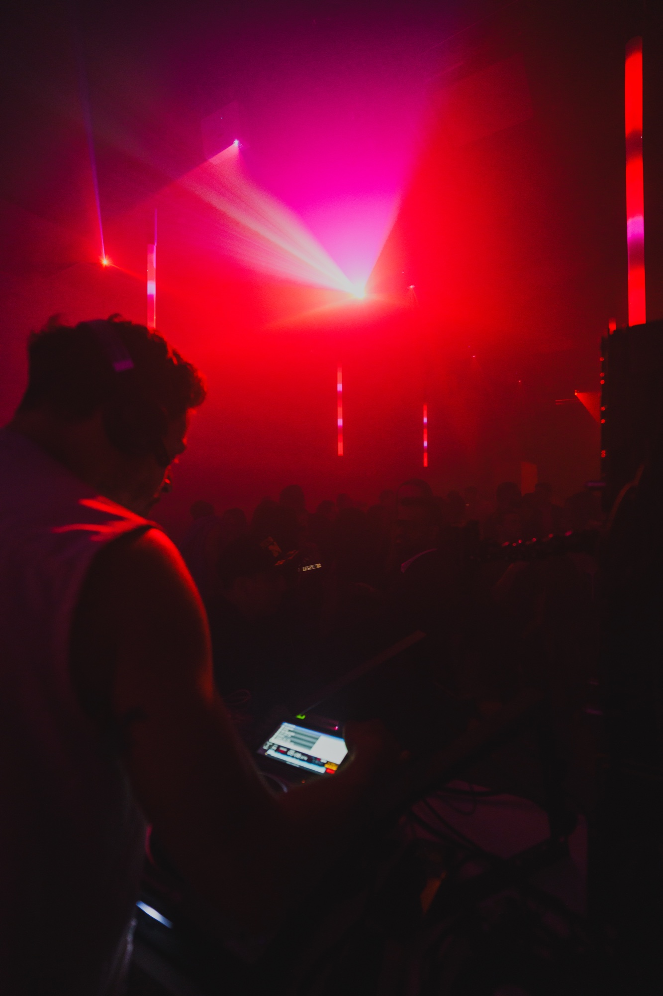 DJ in headphones seen from behind facing a crowd under pink and red beams and haze