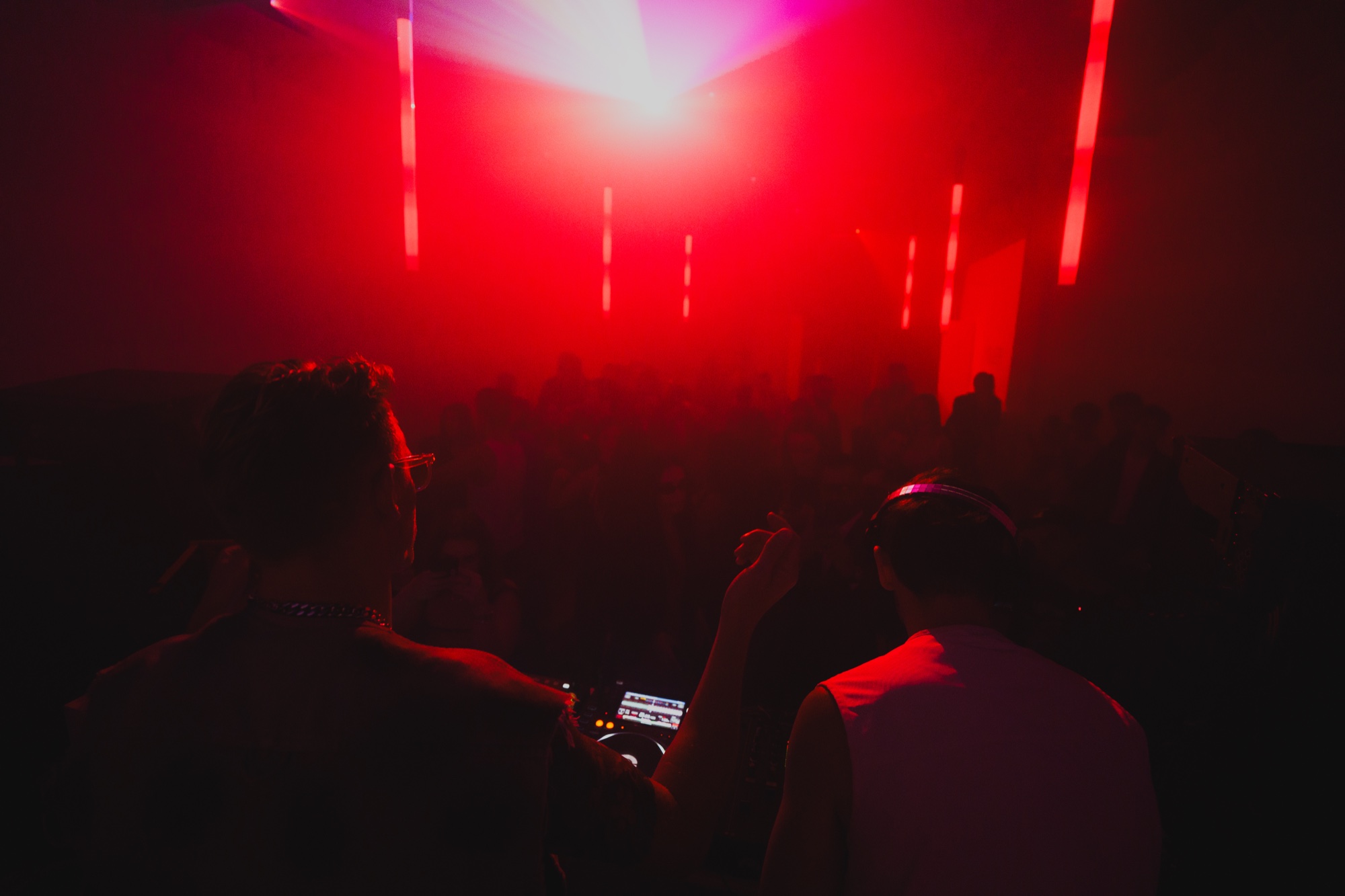 Silhouetted partygoers face a bright red spotlight and tall red light bars through thick haze