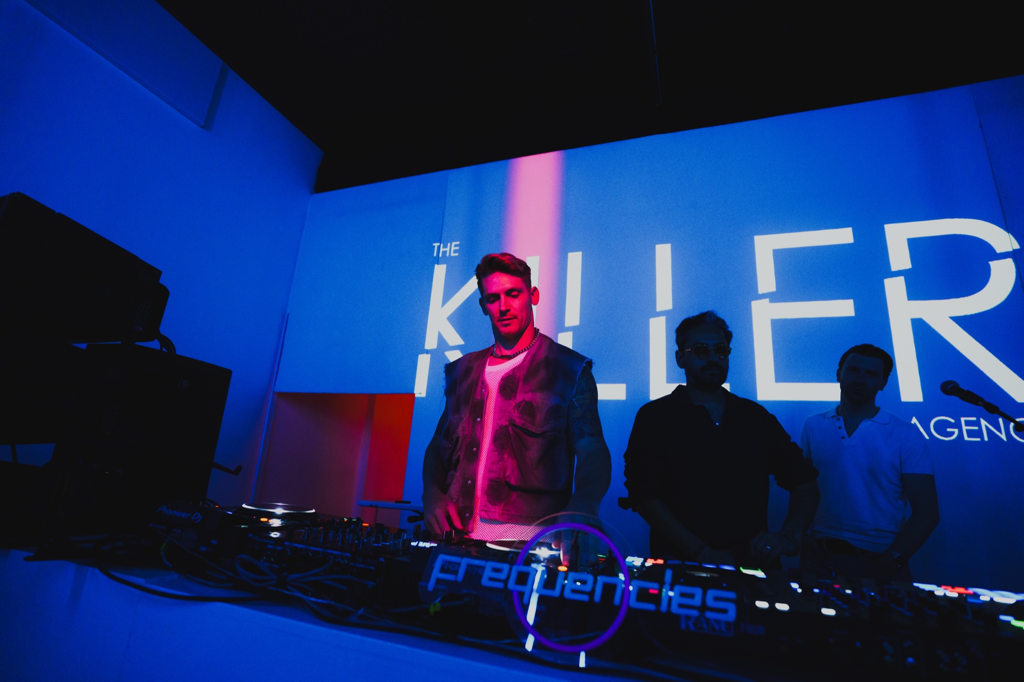 Three men behind DJ decks bathed in blue light beneath The Killer Agency projection at a NYC event